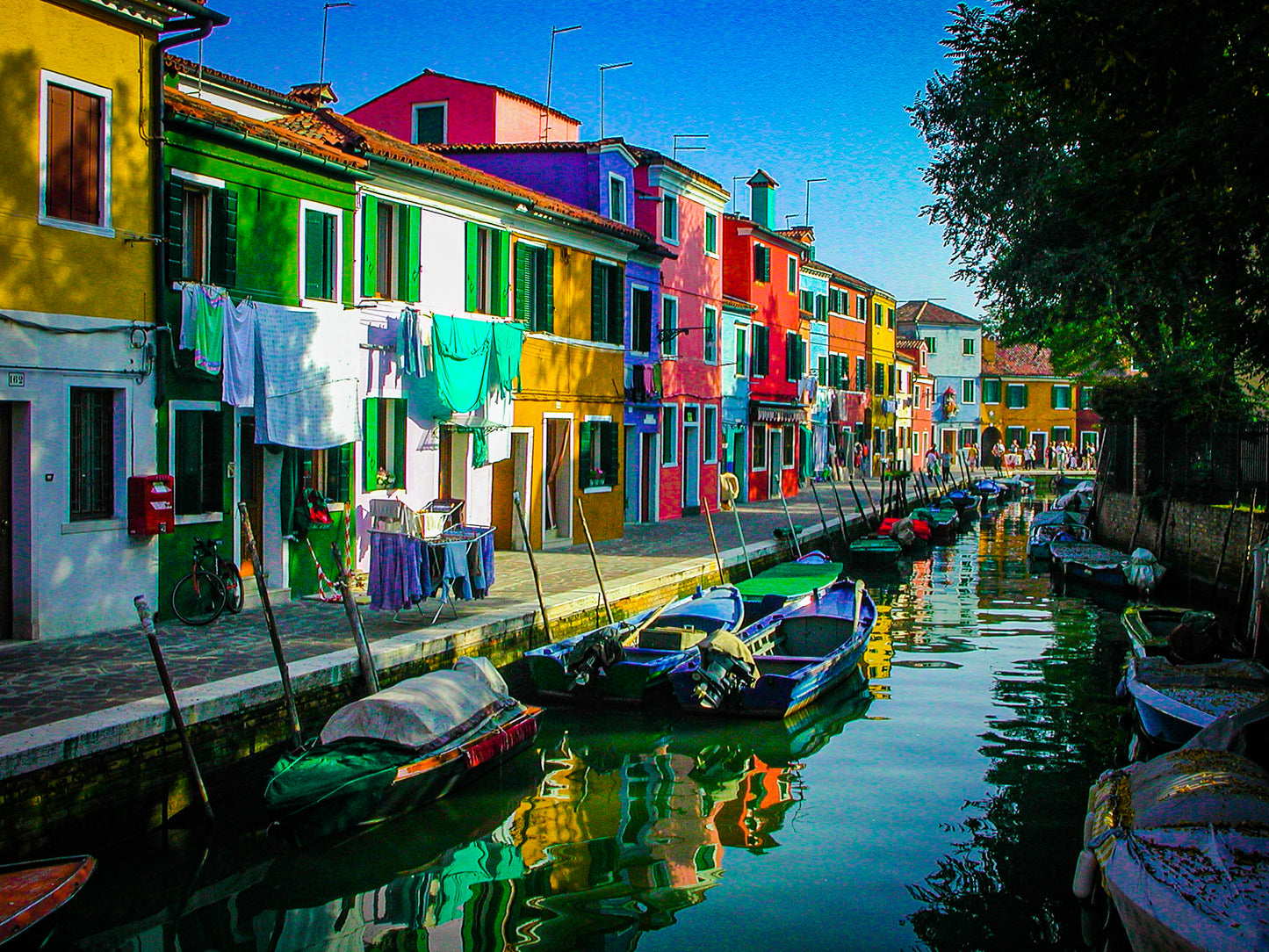 Colorful Burano - Fine Art Print captures the charm of Burano with vibrant houses lining a canal, small boats moored nearby, and laundry hanging from windows in bright sunlight—perfect for lovers of Burano photography.