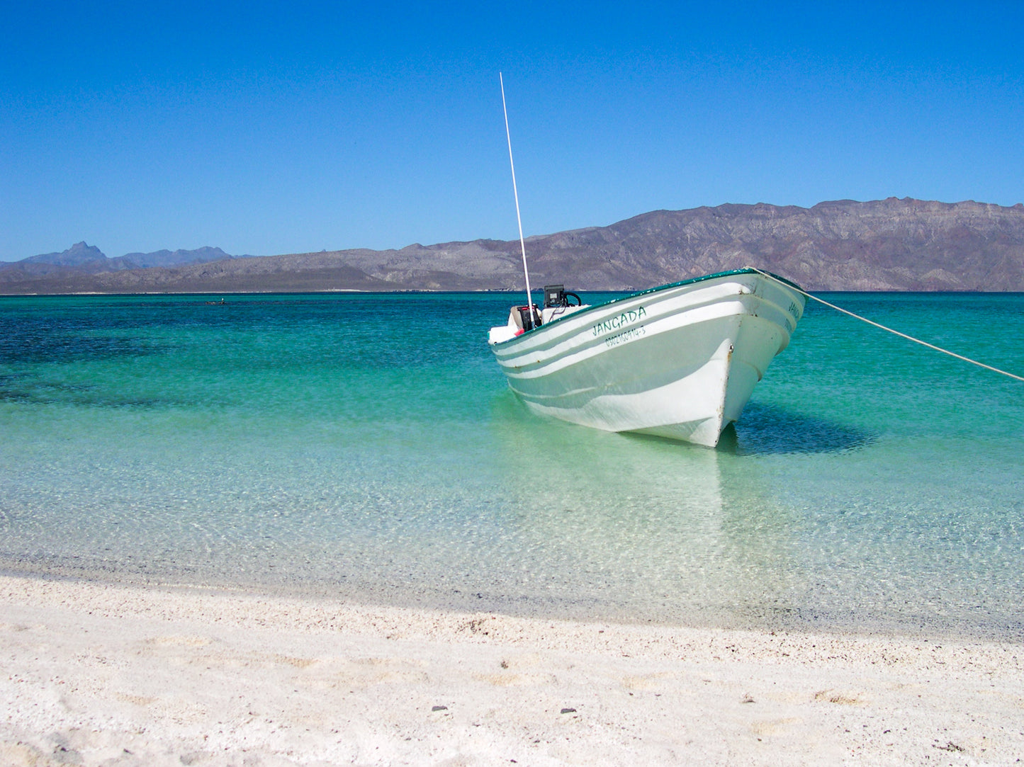 Peaceful Boat - Fine Art Print by Leslie Brashear Photography captures a small white boat in clear turquoise water near a sandy beach, mountains in the distance, and a serene sky—a minimalist Loreto, Mexico seascape.
