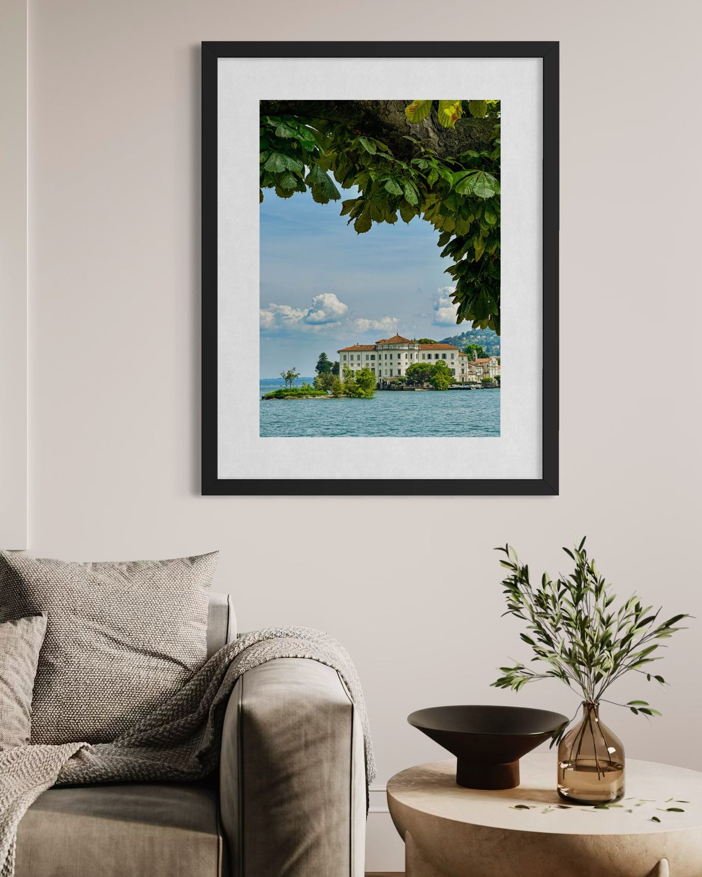 Lago di Maggiore Vista" fine art print by Leslie Brashear Photography hangs on a beige wall above a gray sofa and small plant-topped table, depicting an Italian building by the lake under tree branches.