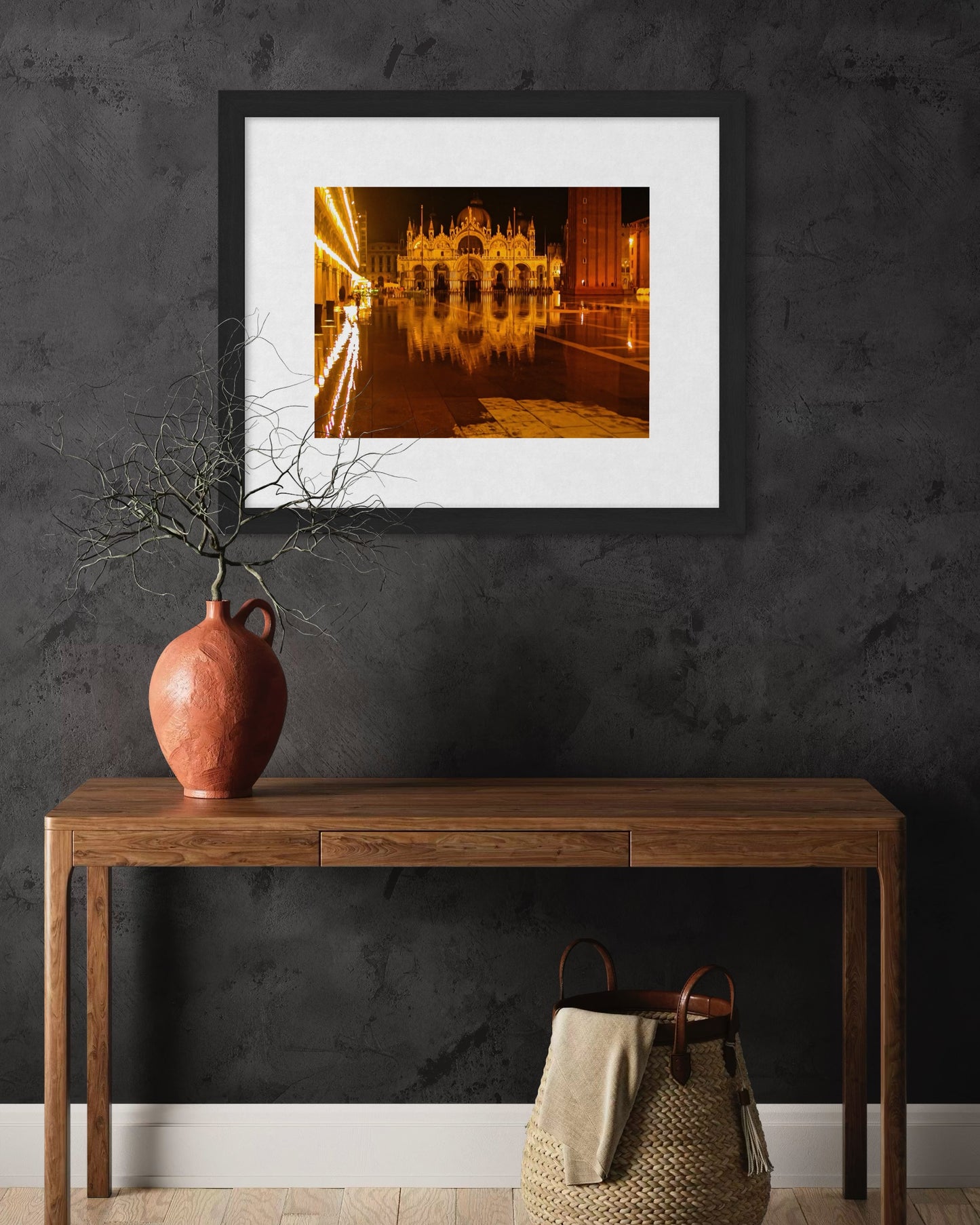 A Flooded St. Marks at Night - Fine Art Print by Leslie Brashear Photography hangs above a wooden table with a terracotta vase, set against a dark wall with a wicker basket underneath.