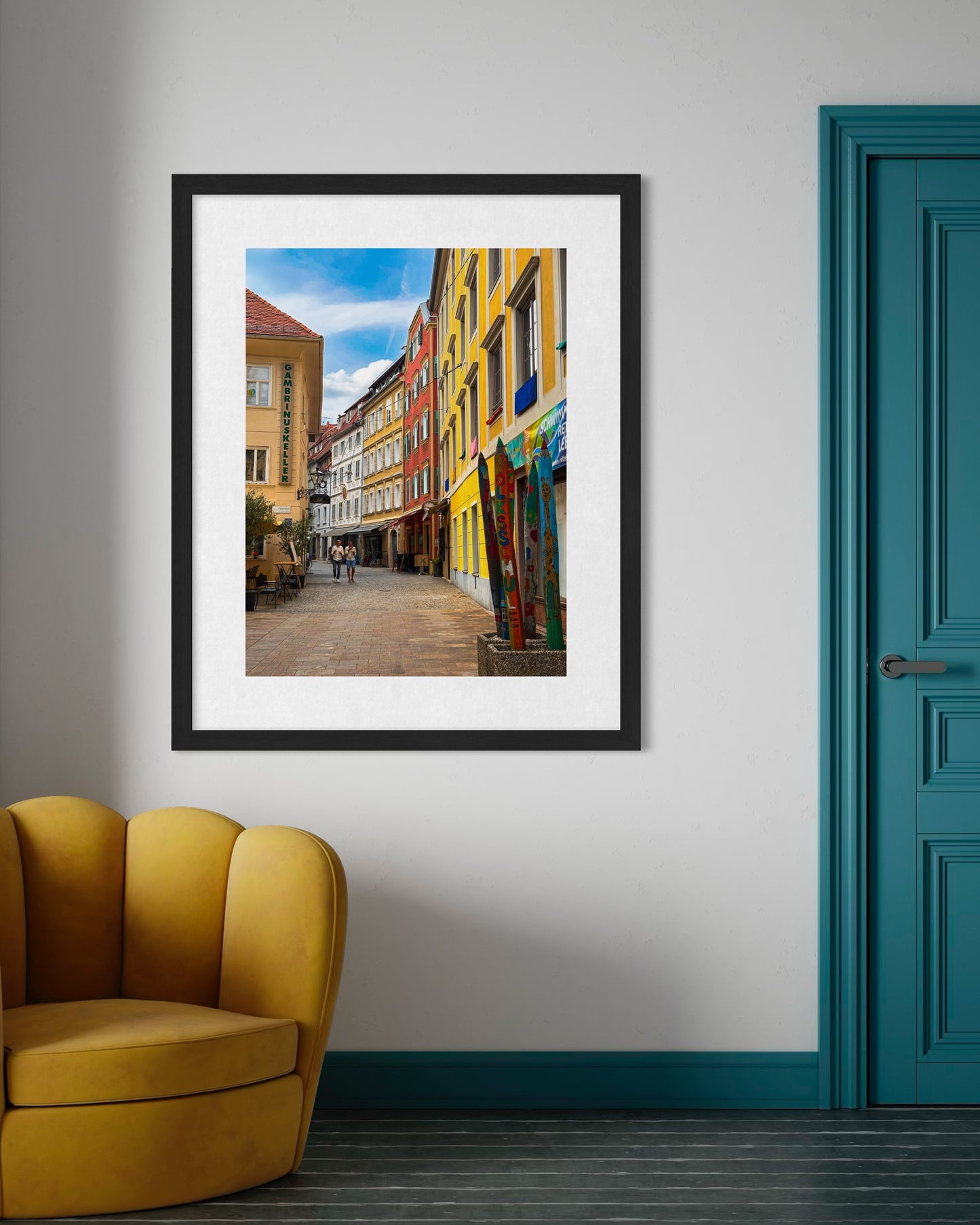 Walking in Graz - Fine Art Print by Leslie Brashear Photography hangs above a mustard yellow chair next to a teal door, showcasing Austria’s vibrant streets and the charm of Graz architecture.
