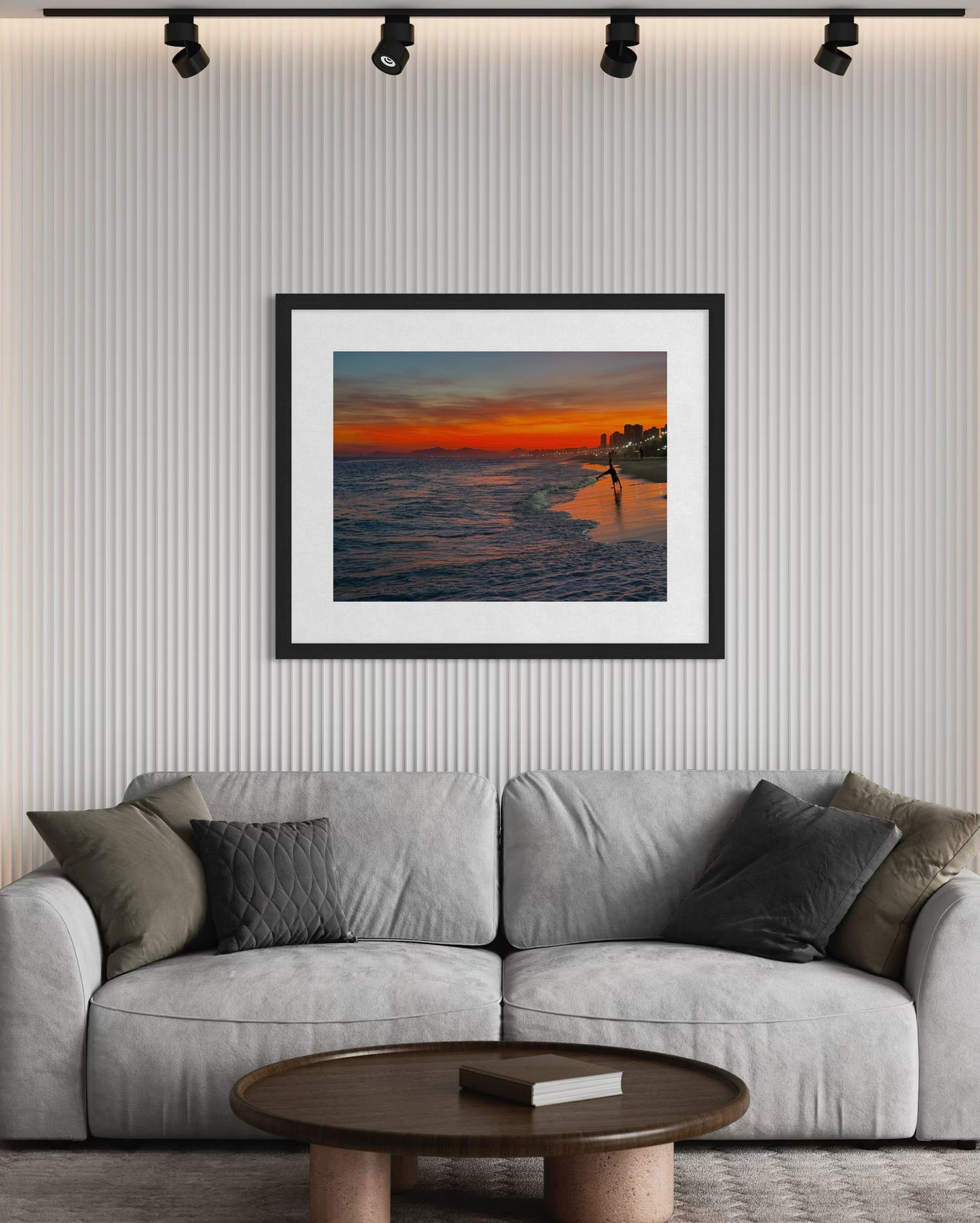 A framed "Sunset Cartwheels at Ipanema Beach - Fine Art Print" by Leslie Brashear Photography hangs on a textured wall above a gray couch with assorted cushions and a round coffee table in front.