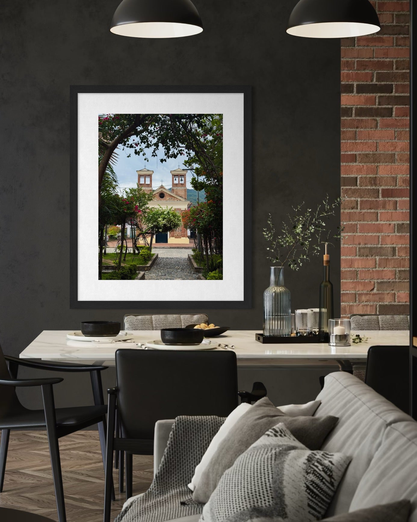 A modern dining room features a white table, black chairs, and “Charming Church - Fine Art Print” by Leslie Brashear Photography, displaying Colombian architecture from Santa Fe de Antioquia on a dark wall.