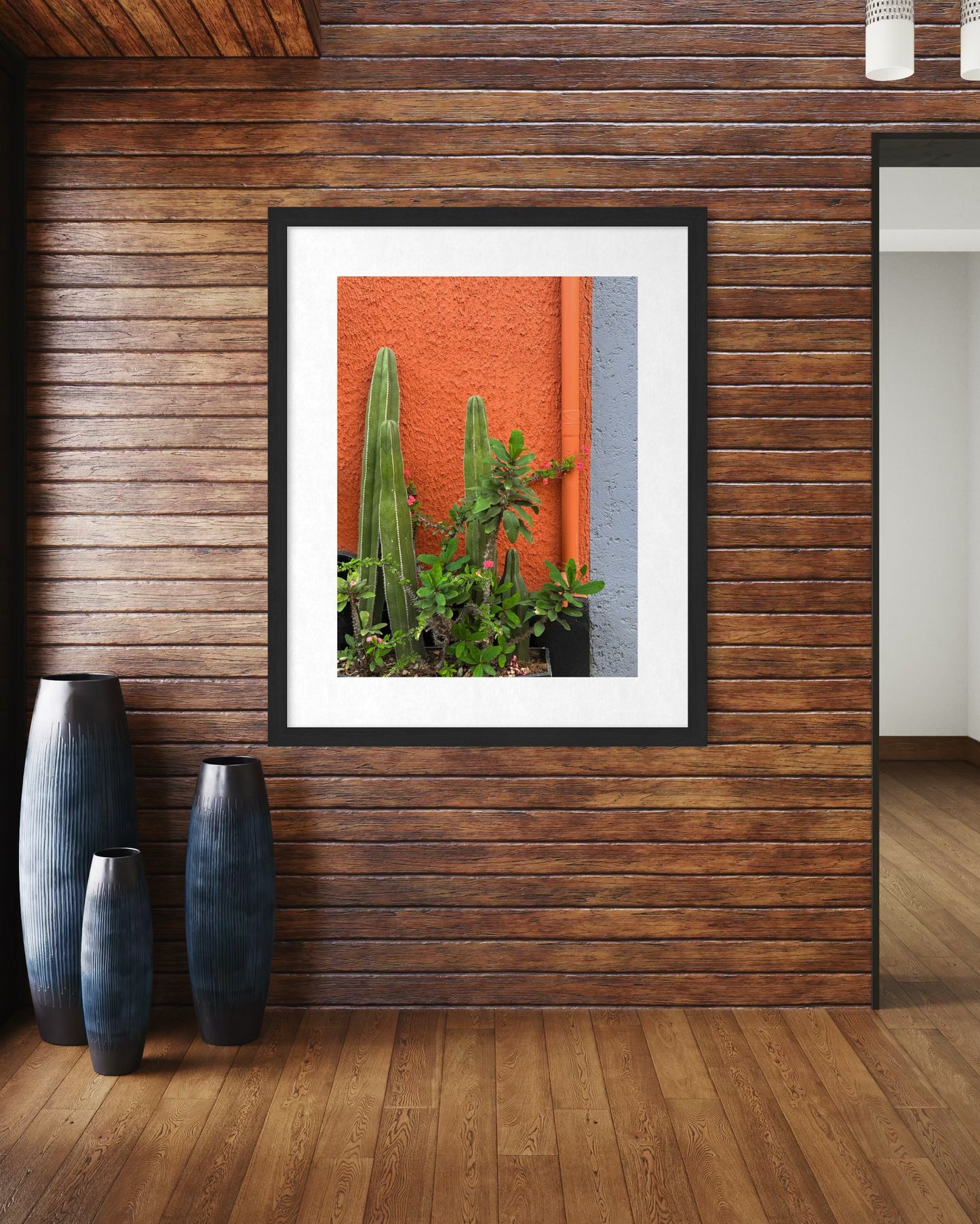 Cactus on Display - Fine Art Print by Leslie Brashear Photography features vibrant green cacti and a succulent, framed and mounted on wooden paneling beside ceramic vases, adding a modern touch to a red and gray wall.