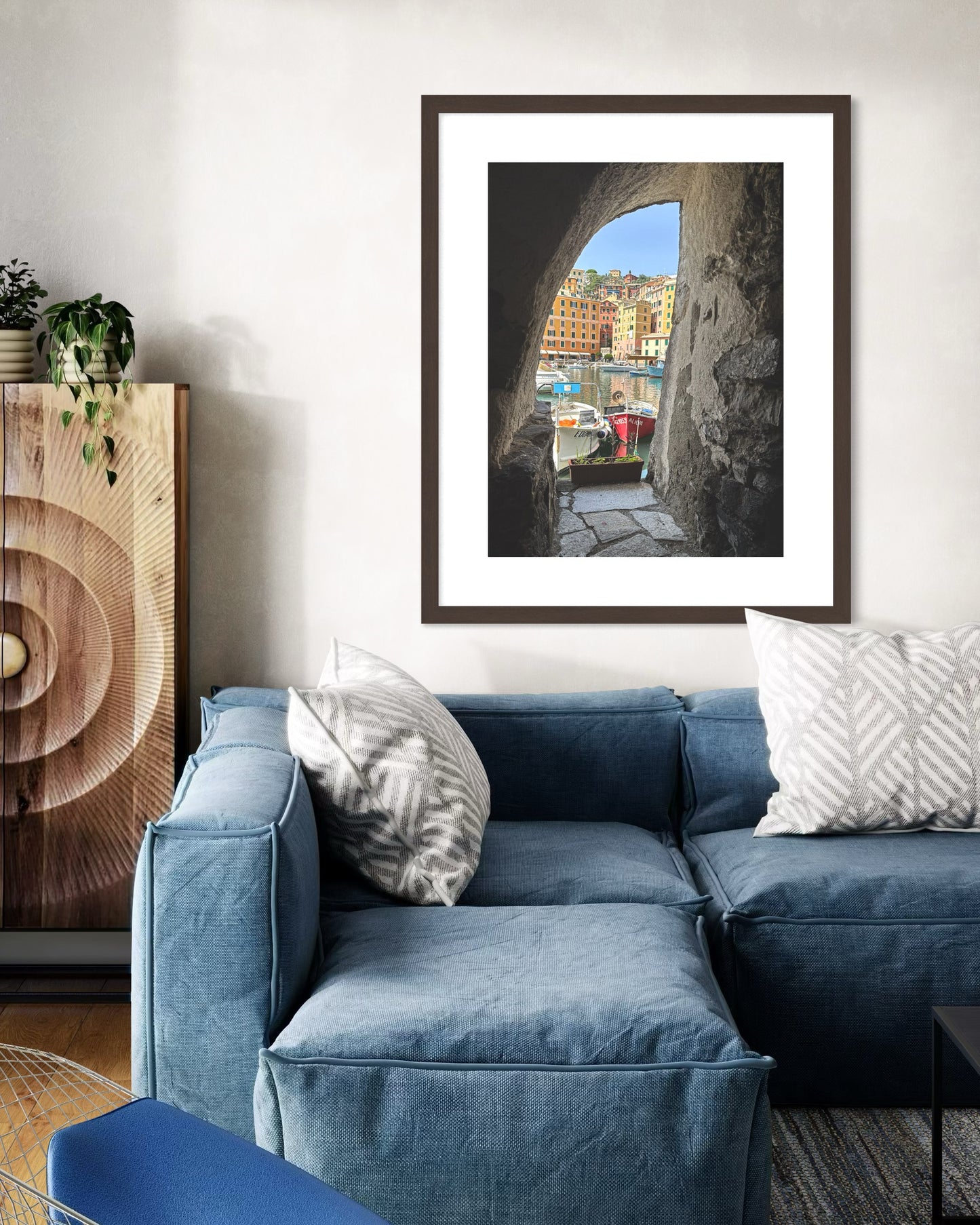 A living room with a blue sofa, patterned pillows, a plant on a wooden cabinet, and Leslie Brashear Photography's "Framed Serenity of Camogli Harbor" fine art print displayed on the wall.