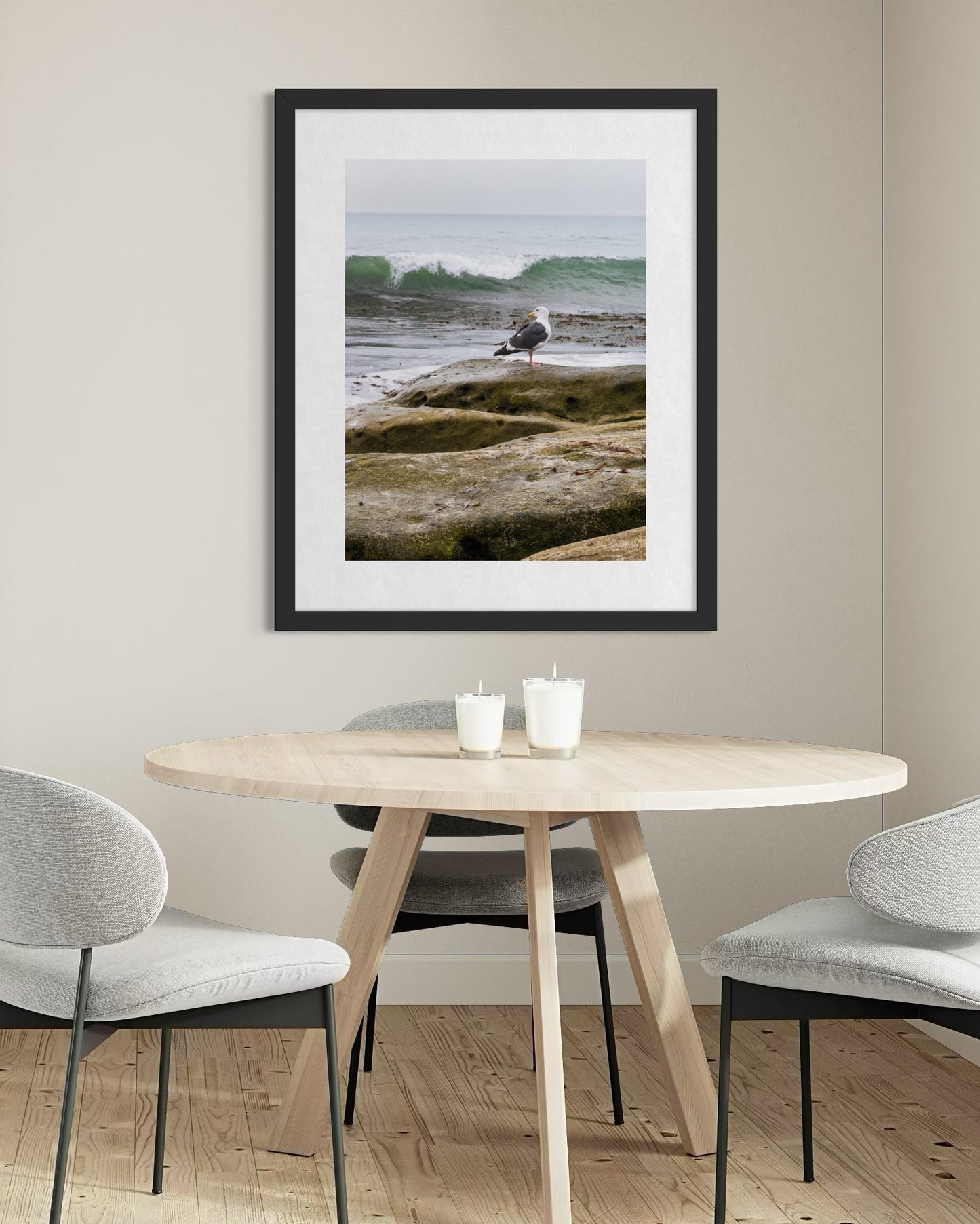 A modern dining room features a round wooden table, three gray chairs, two candles, and "Seagull on the Rocky Shoreline - Fine Art Print" by Leslie Brashear Photography—adding an elegant touch of coastal artistry to the wall.