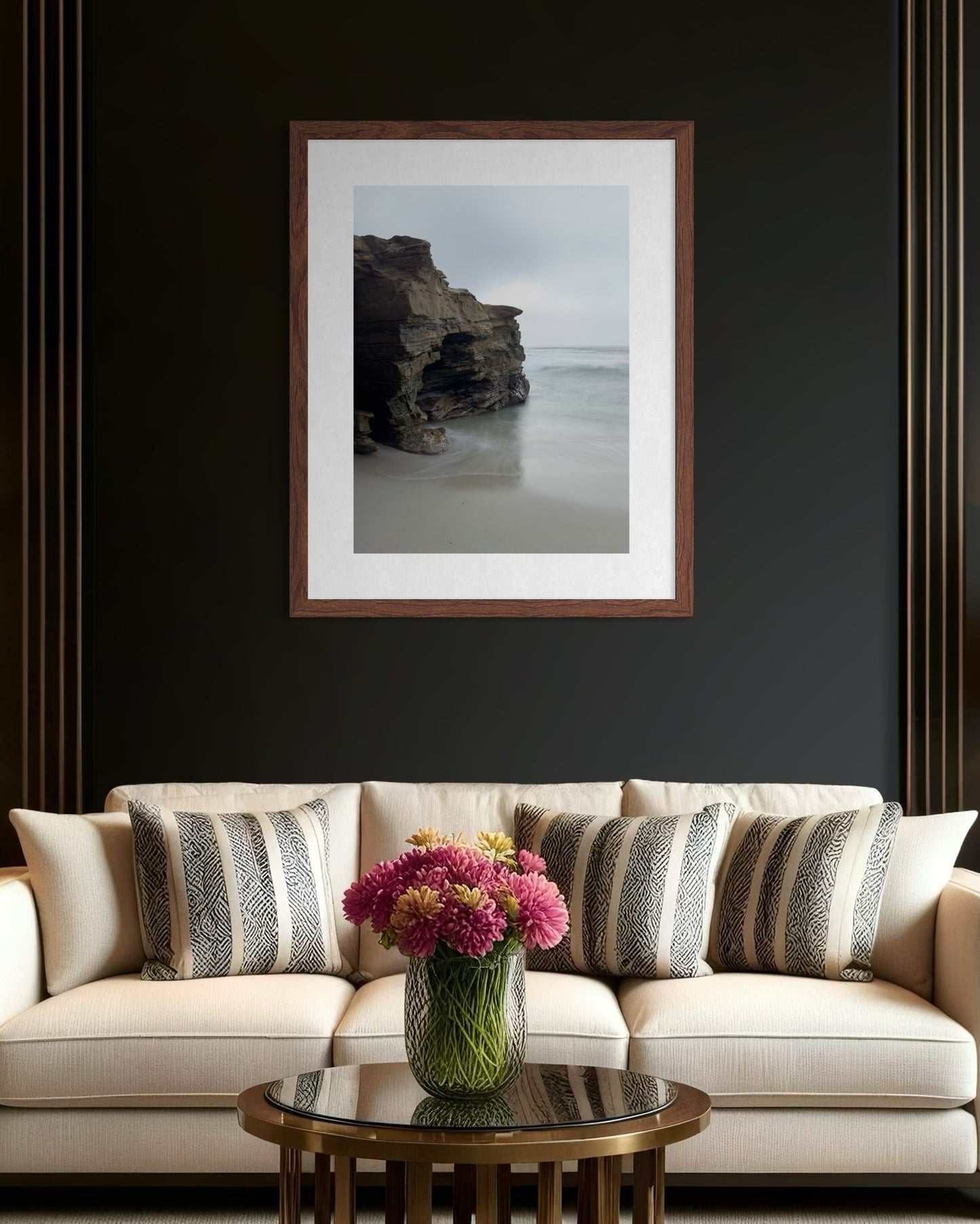 A beige sofa with patterned cushions sits against a dark wall, above which hangs "Rugged Coastal Cliffs at Windansea - Fine Art Print" by Leslie Brashear Photography. A round table with a vase of flowers is placed in front of the sofa.