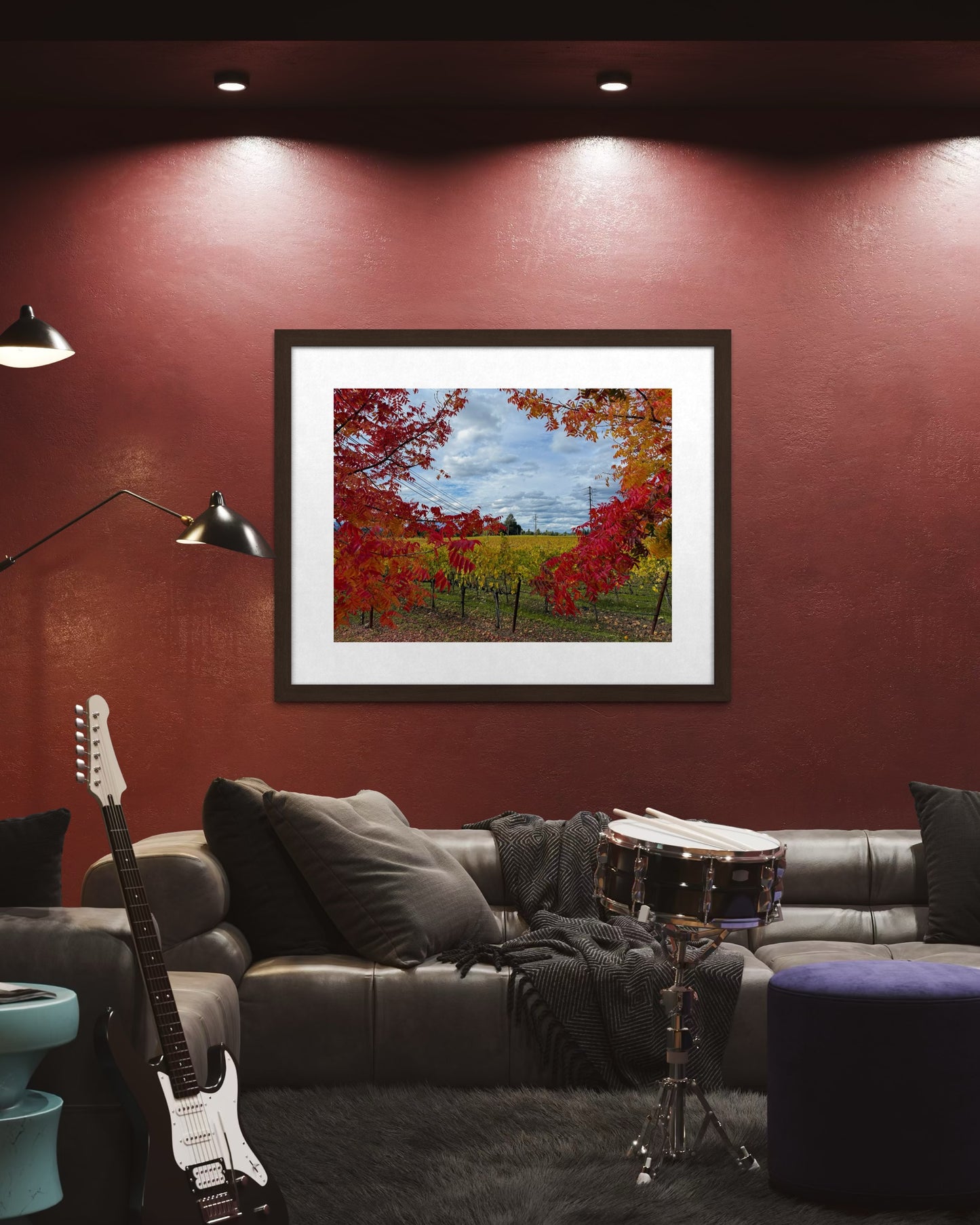 A modern living room with a gray sofa, drum, and electric guitar. On the dark red wall above the sofa hangs "Autumn Vineyard in Napa Valley - Fine Art Print" by Leslie Brashear Photography, showcasing vivid red foliage.