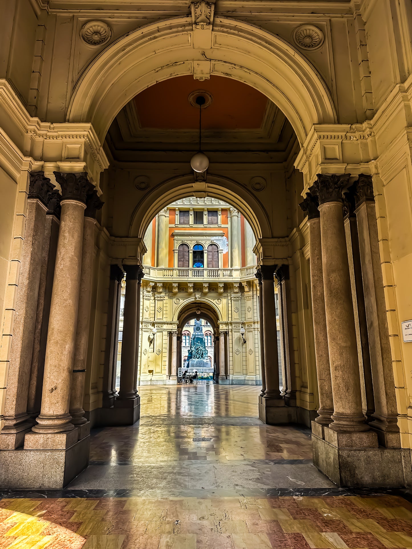 The "Timeless Arches of Pavia - Fine Art Print" by Leslie Brashear Photography captures a grand archway with historic columns, leading to a courtyard centered around a fountain and surrounded by ornate Italian architecture.