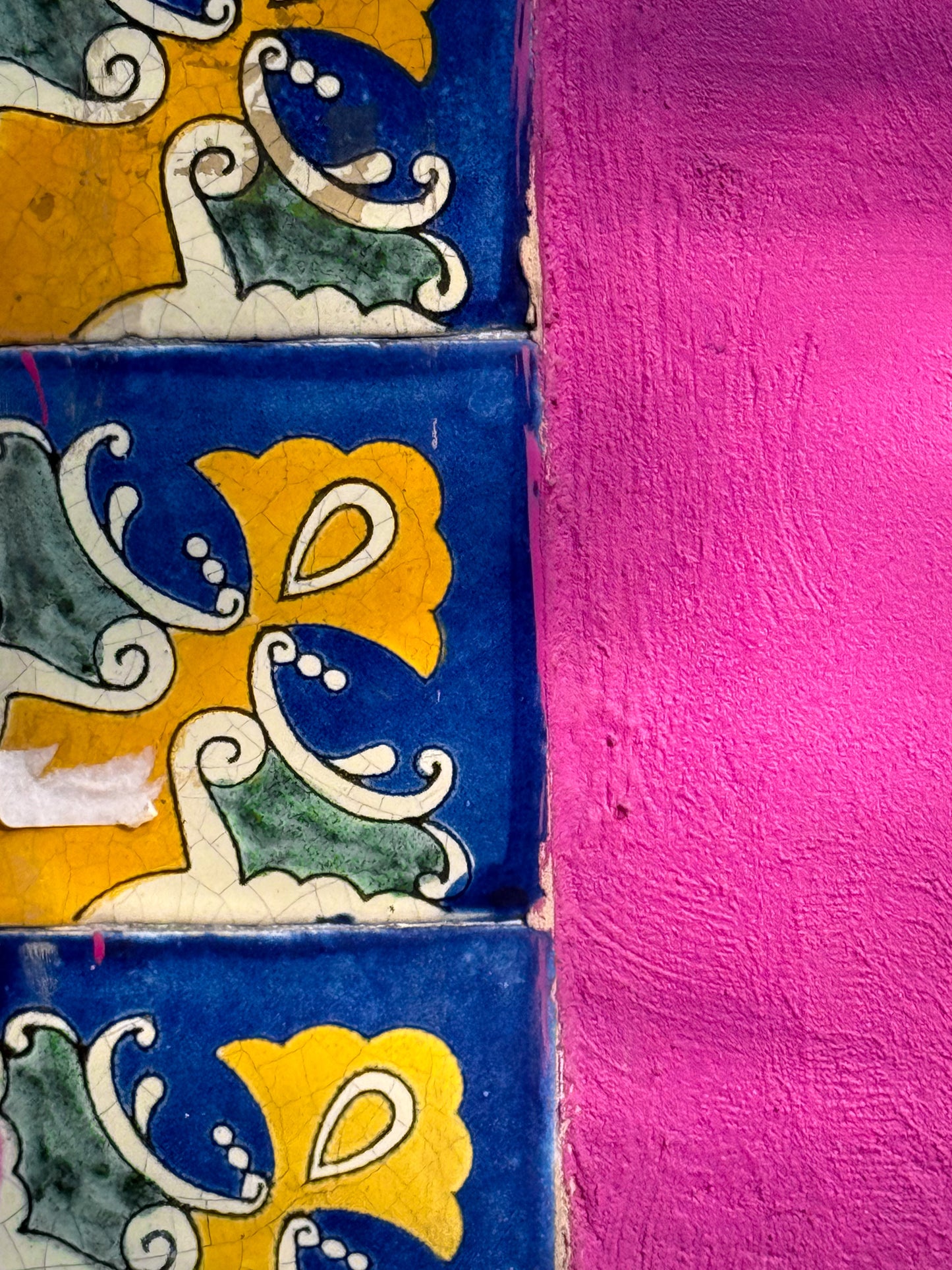 The "Vibrant Tiles and Bright Pink Wall - Fine Art Print" by Leslie Brashear Photography showcases three floral tiles in blue, yellow, and green beside a textured, vivid pink stucco wall.