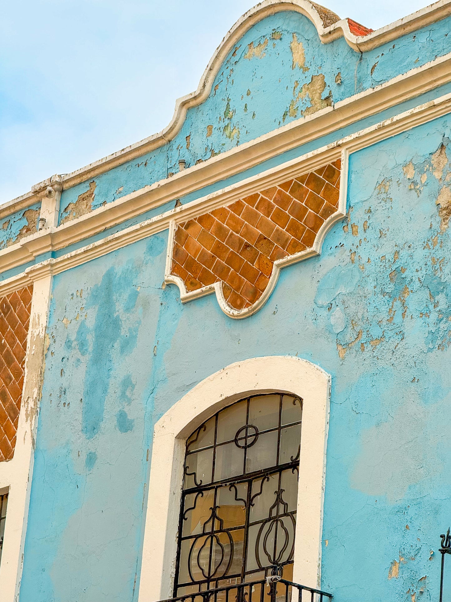 Blue Facade in Puebla - Fine Art Print by Leslie Brashear Photography captures a weathered blue building with peeling paint, an arched window with ironwork, and brown geometric accents—an embodiment of Mexican facade art.