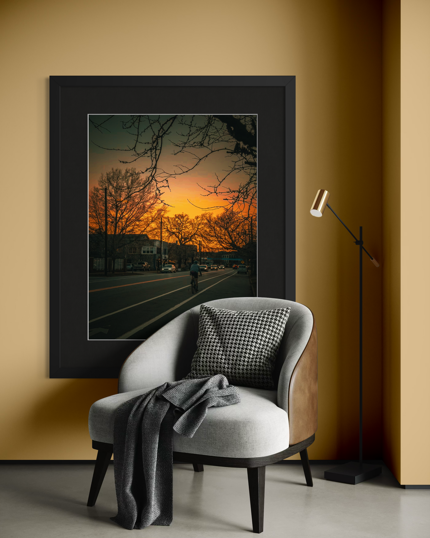 A modern armchair with a blanket and cushion sits before "Cycling at Sunset" by Leslie Brashear Photography, a large framed photo of evening cycling, beside a slim black floor lamp against a beige wall.