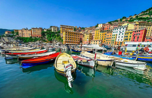 Colorful boats float in a calm harbor of Camogli, Italy, with vibrant buildings lining the hillside under a clear blue sky.