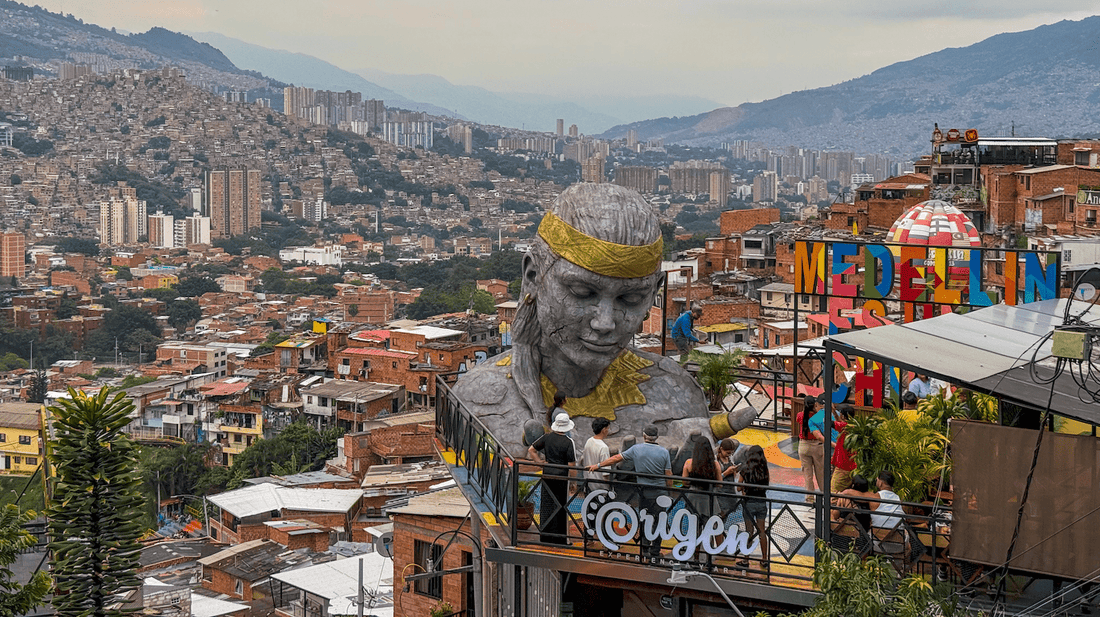Large stone sculpture of a head overlooks a cityscape with hills, buildings, and a colorful sign reading "Medellin" in the foreground. People are gathered on a rooftop terrace.