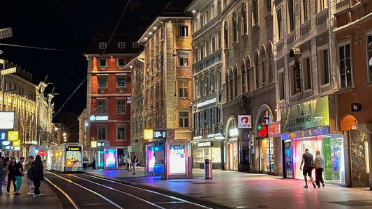 A city street in Graz at night with illuminated shop signs, tram tracks, and pedestrians walking along the sidewalks.