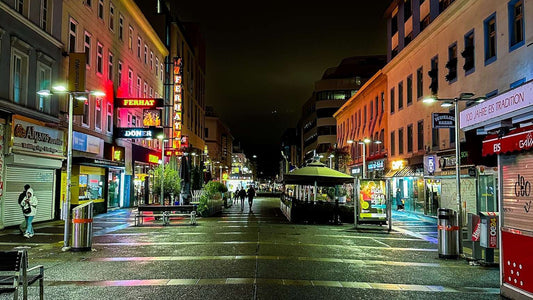 A wide pedestrian street in Vienna at night is lined with brightly lit shops and restaurants, with a few people walking and benches along the center.