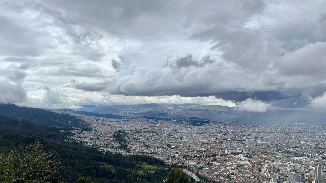 Aerial view of a densely built cityscape, Bogota Colombia, surrounded by mountains under a cloudy sky. The scene shows urban and green areas merging at the city's edges.
