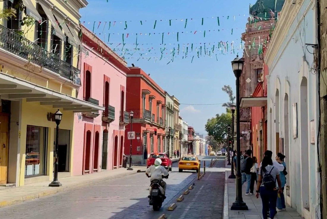 A vibrant street scene in Oaxaca features colorful buildings, hanging decorations, pedestrians, a motorcyclist, and a yellow taxi—capturing the lively spirit and everyday adventure in Oaxaca on a sunny day.