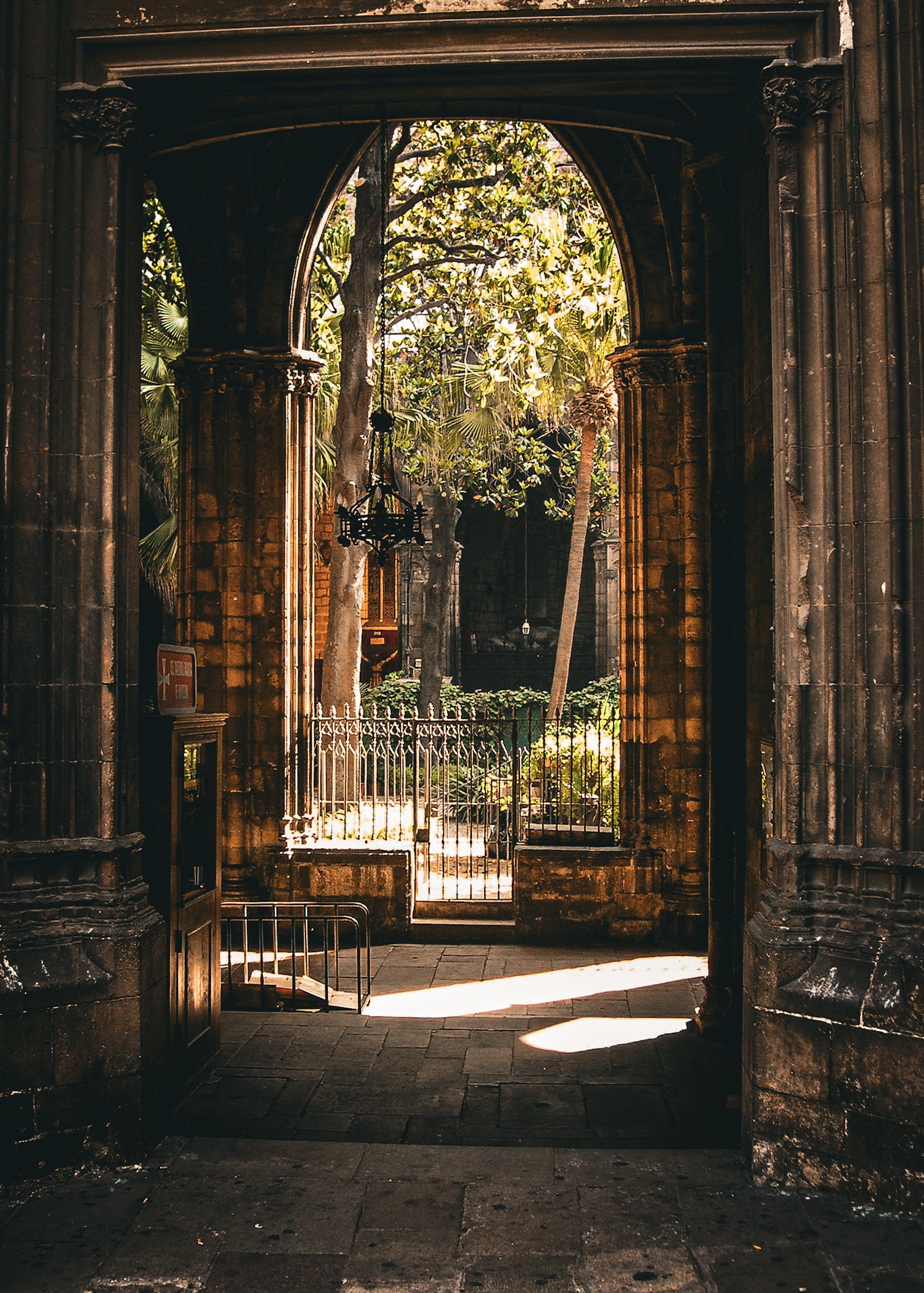 The Barcelona Arch - Fine Art Print features an arched stone doorway leading to a sunlit courtyard with trees, plants, and an iron fence, where shadows and light dance throughout the scene.