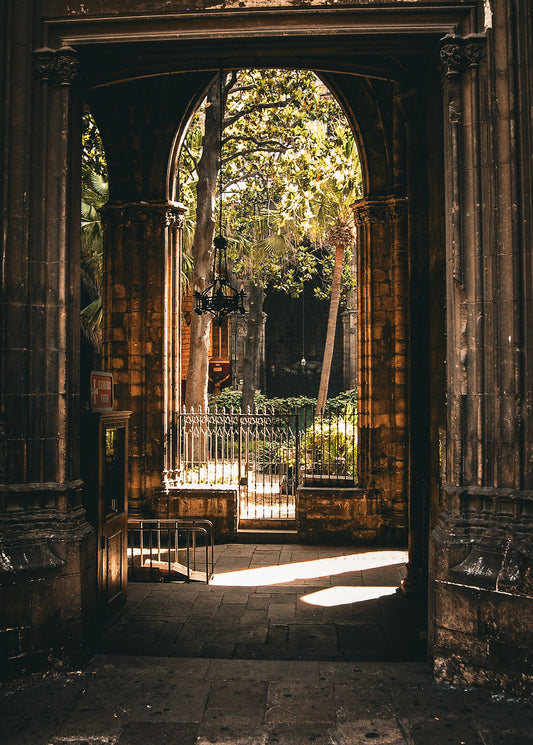 The Barcelona Arch - Fine Art Print features an arched stone doorway leading to a sunlit courtyard with trees, plants, and an iron fence, where shadows and light dance throughout the scene.