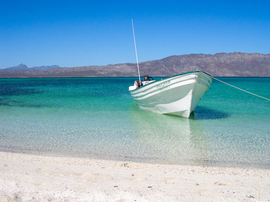 Peaceful Boat - Fine Art Print by Leslie Brashear Photography captures a small white boat in clear turquoise water near a sandy beach, mountains in the distance, and a serene sky—a minimalist Loreto, Mexico seascape.