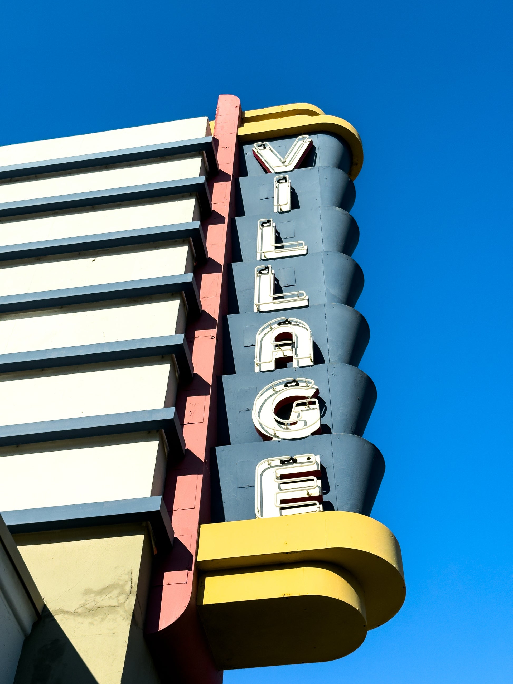 Village Theater on Coronado Island - Fine Art Print by Leslie Brashear Photography features an art deco sign with "VILLAGE" in white letters on blue, capturing the iconic Coronado theater against a clear blue sky.
