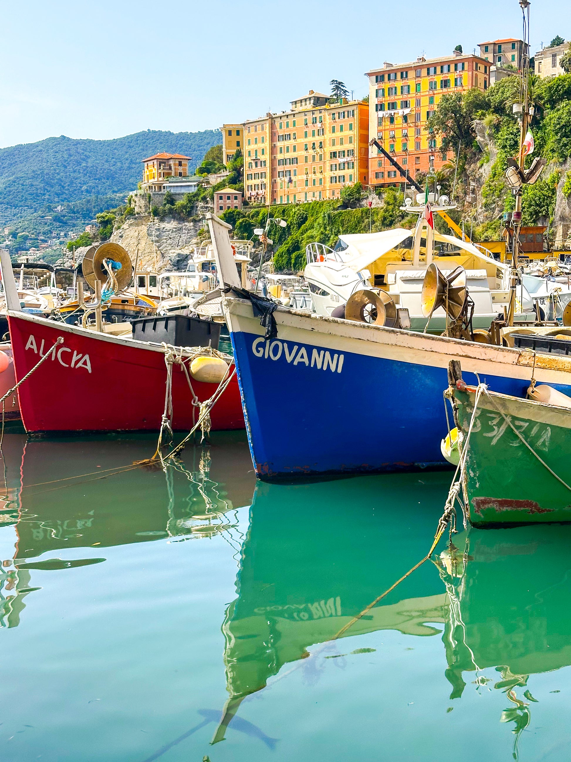 Reflections - Fine Art Print by Leslie Brashear Photography captures vibrant fishing boats in Camogli harbor, Italy, set against multistory yellow and orange hillside buildings beneath a clear sky.