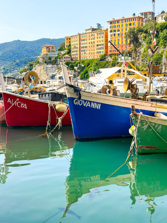 Reflections - Fine Art Print by Leslie Brashear Photography captures vibrant fishing boats in Camogli harbor, Italy, set against multistory yellow and orange hillside buildings beneath a clear sky.