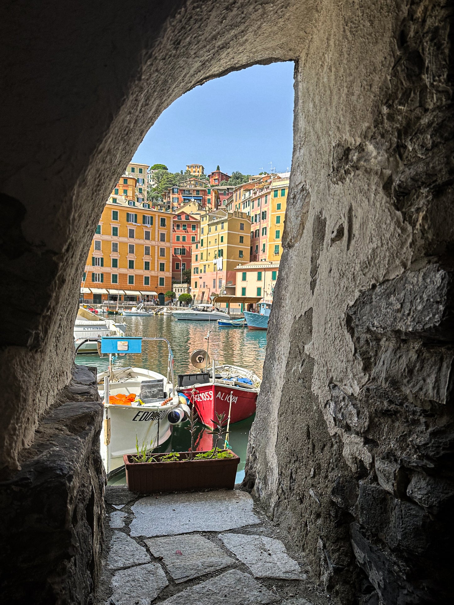 Framed Serenity of Camogli Harbor – Fine Art Print by Leslie Brashear Photography captures boats docked in a harbor, colorful buildings, and hillside through a stone archway—a timeless view of this charming Italian coastal town.
