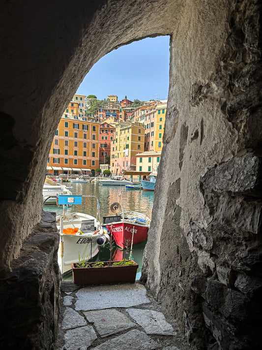 Framed Serenity of Camogli Harbor – Fine Art Print by Leslie Brashear Photography captures boats docked in a harbor, colorful buildings, and hillside through a stone archway—a timeless view of this charming Italian coastal town.