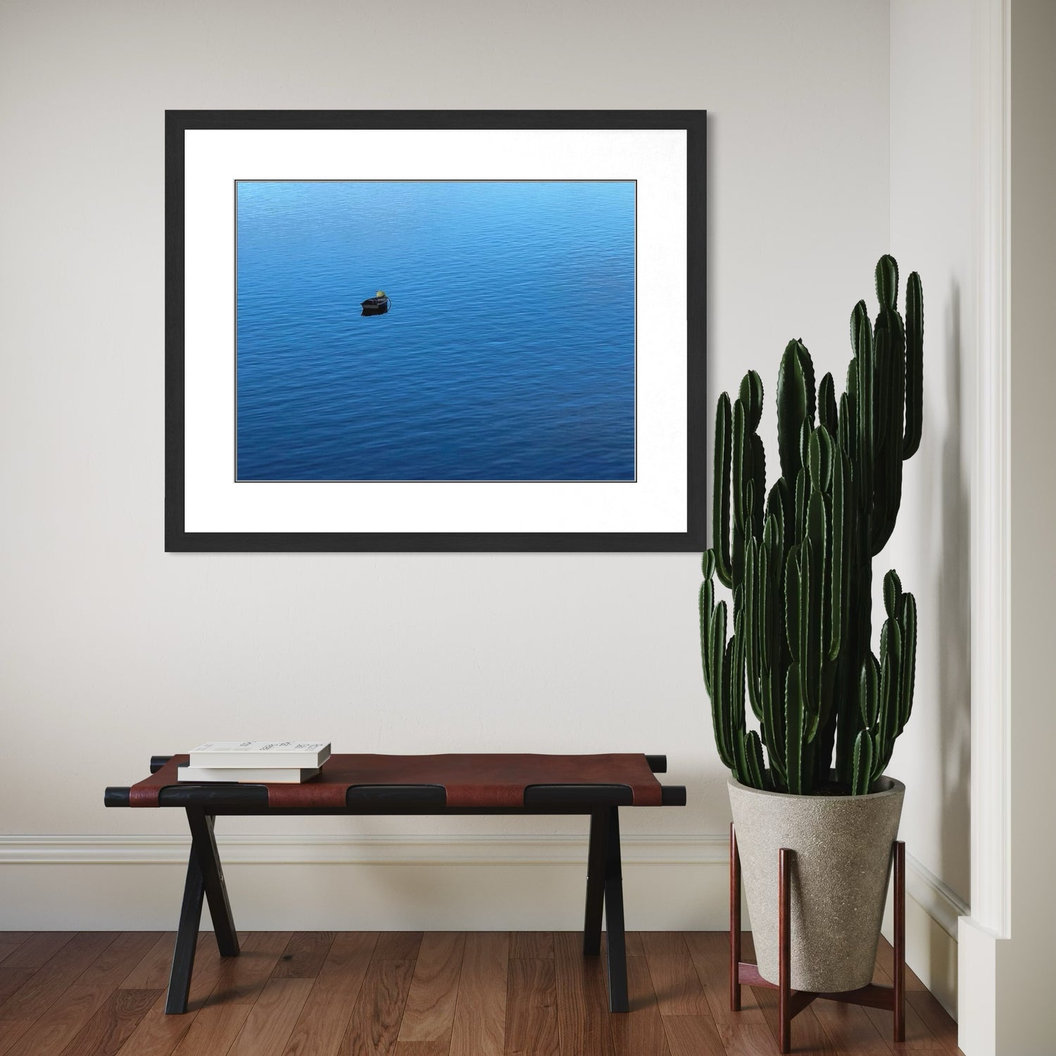 Framed artwork of a blue landscape with a small boat, placed above a wooden bench with a cactus plant beside it.