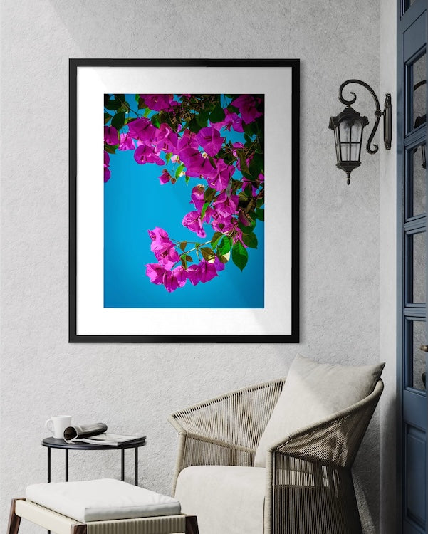 The Bougainvillea - Fine Art Print by Leslie Brashear Photography features vibrant pink bougainvillea blooms against a blue sky, making it perfect tropical wall art above a wicker chair and small table.