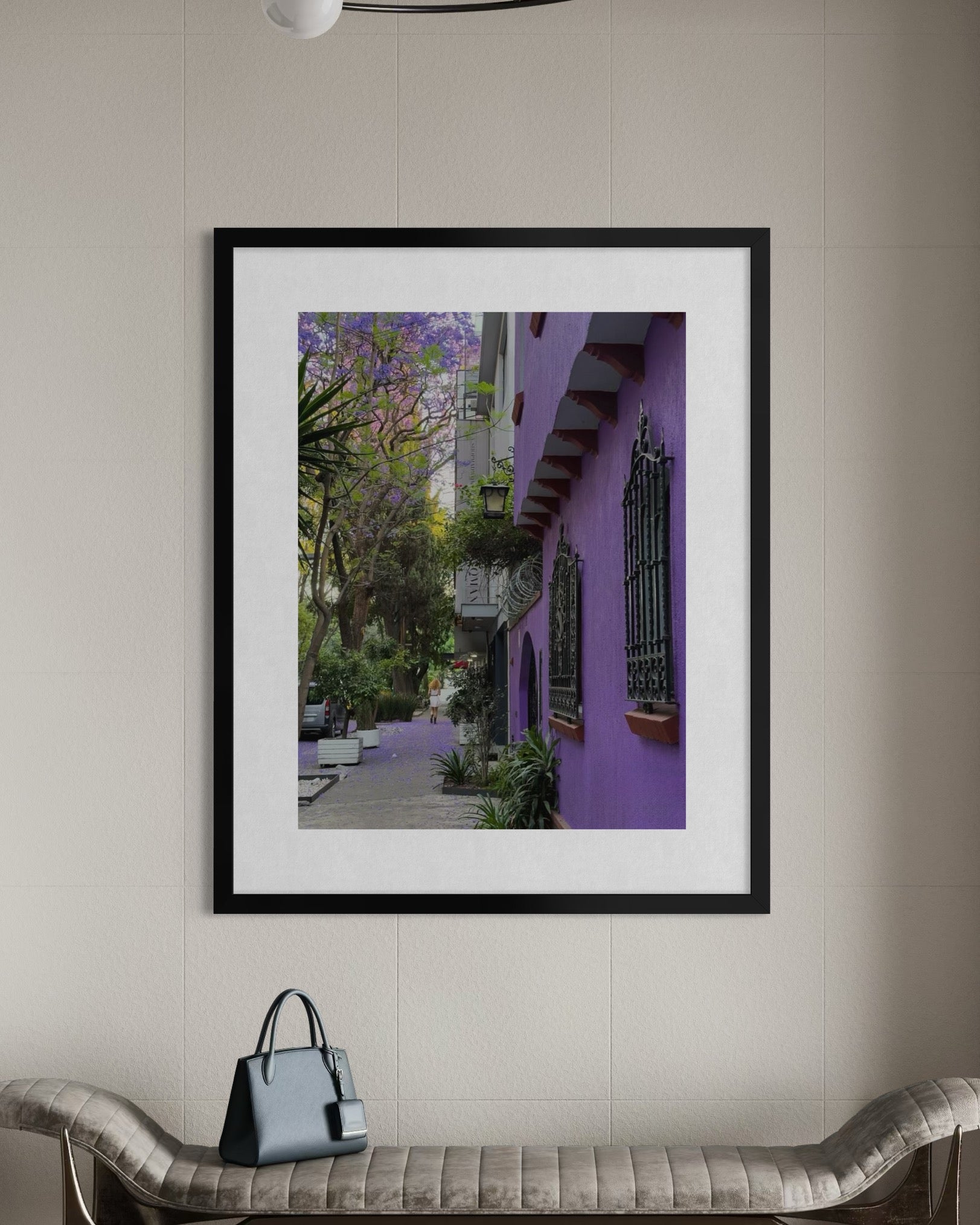 Purple Streetscape - Fine Art Print by Leslie Brashear Photography shows a purple building with iron grilles and jacaranda tree, framed and displayed on a beige wall above a bench holding a gray handbag.