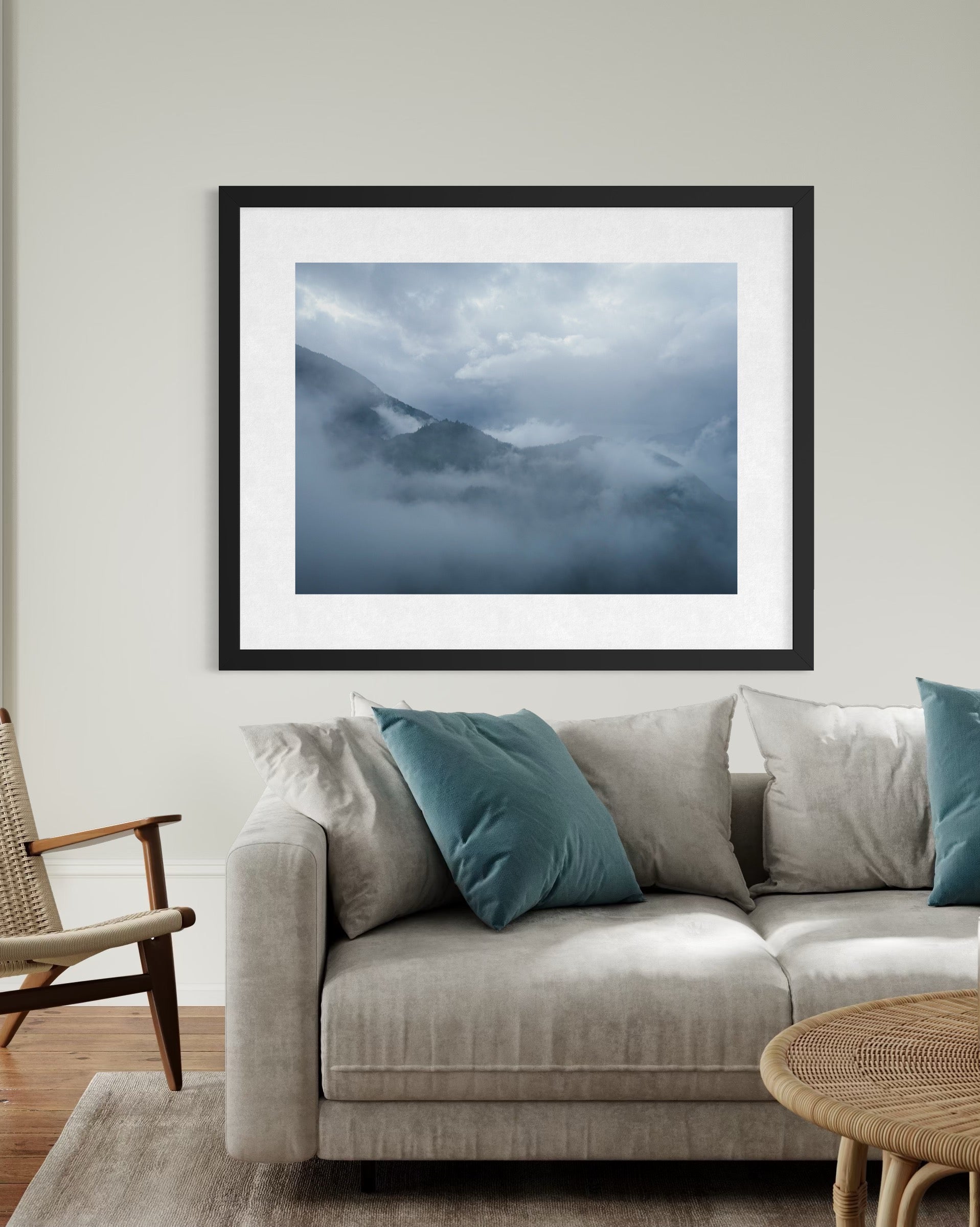 A Misty Mountain View - Fine Art Print by Leslie Brashear Photography is displayed above a beige sofa with blue and gray pillows, adding a modern touch to the living room’s light-colored wall.