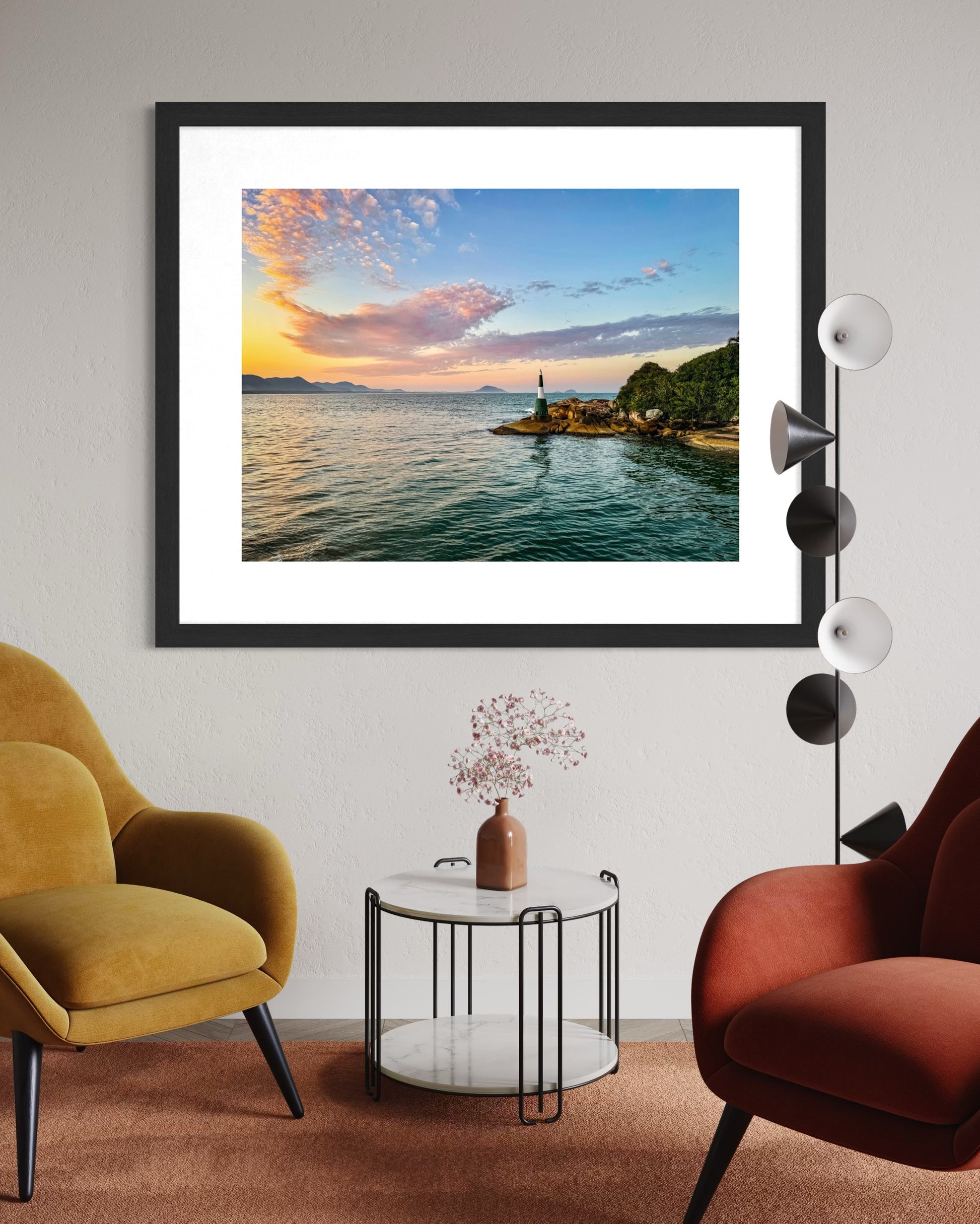 A modern living room with mustard and rust chairs, a round table, and Coastal Sunset - Fine Art Print by Leslie Brashear Photography featuring the Florianópolis lighthouse on the wall.