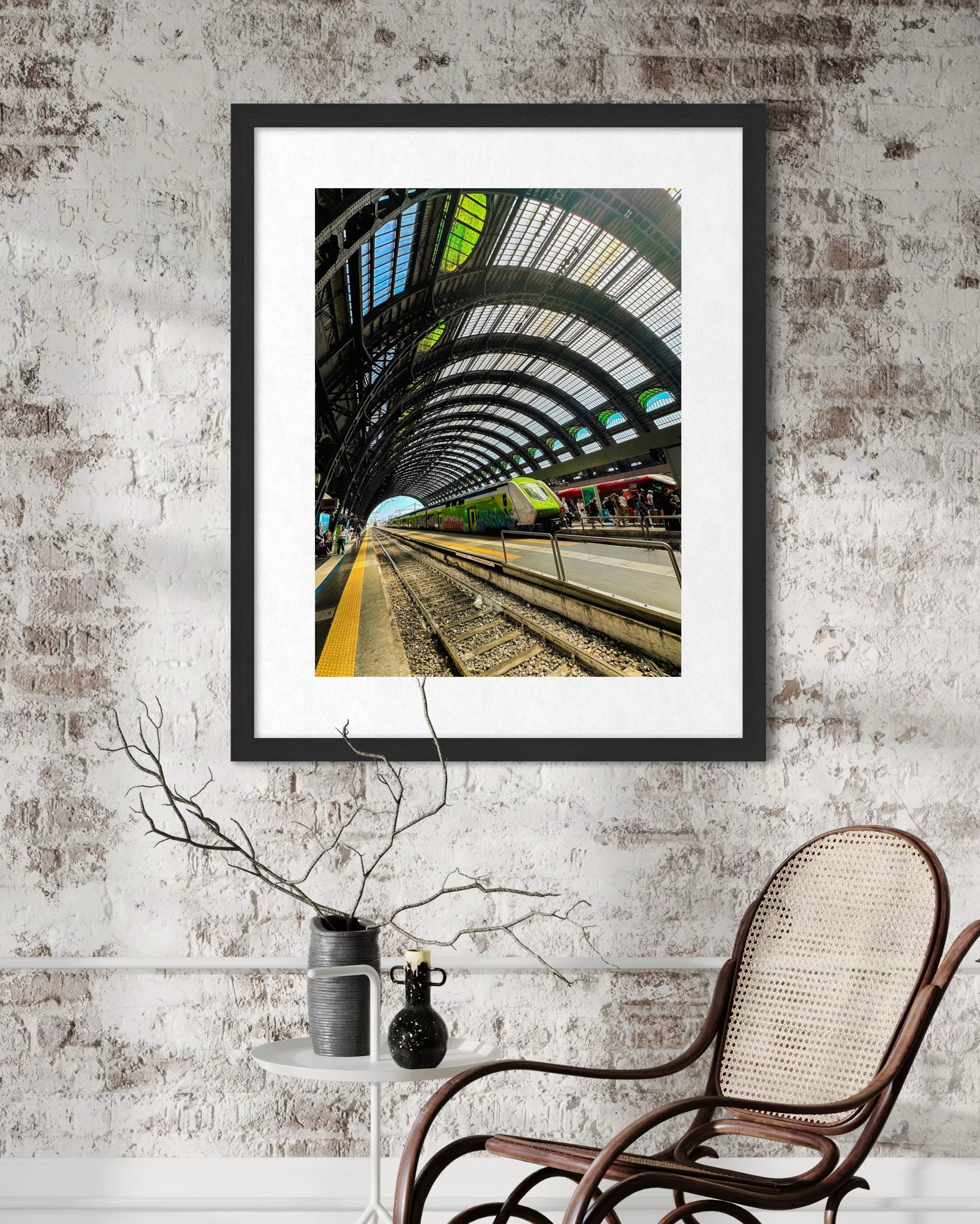 A Milano Centrale - Fine Art Print by Leslie Brashear Photography, featuring the station's iconic arched glass roof, is displayed on a textured wall above a small round table and wicker chair.