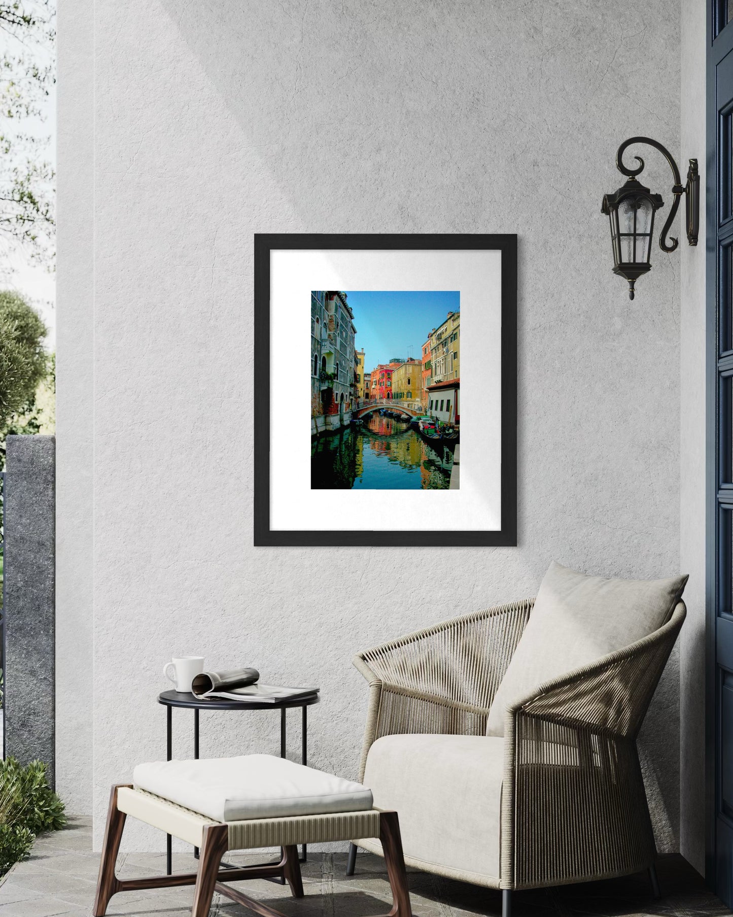 A Quiet Venice Canal - Fine Art Print by Leslie Brashear Photography is framed and hangs on a light gray wall above a wicker chair with cushions, beside a small round table and a wall-mounted lantern.