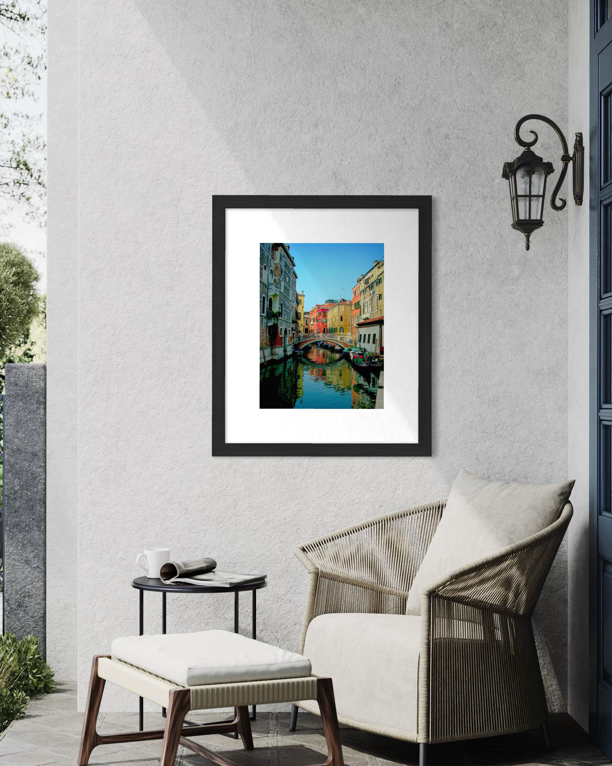 A Quiet Venice Canal - Fine Art Print by Leslie Brashear Photography is framed and hangs on a light gray wall above a wicker chair with cushions, beside a small round table and a wall-mounted lantern.