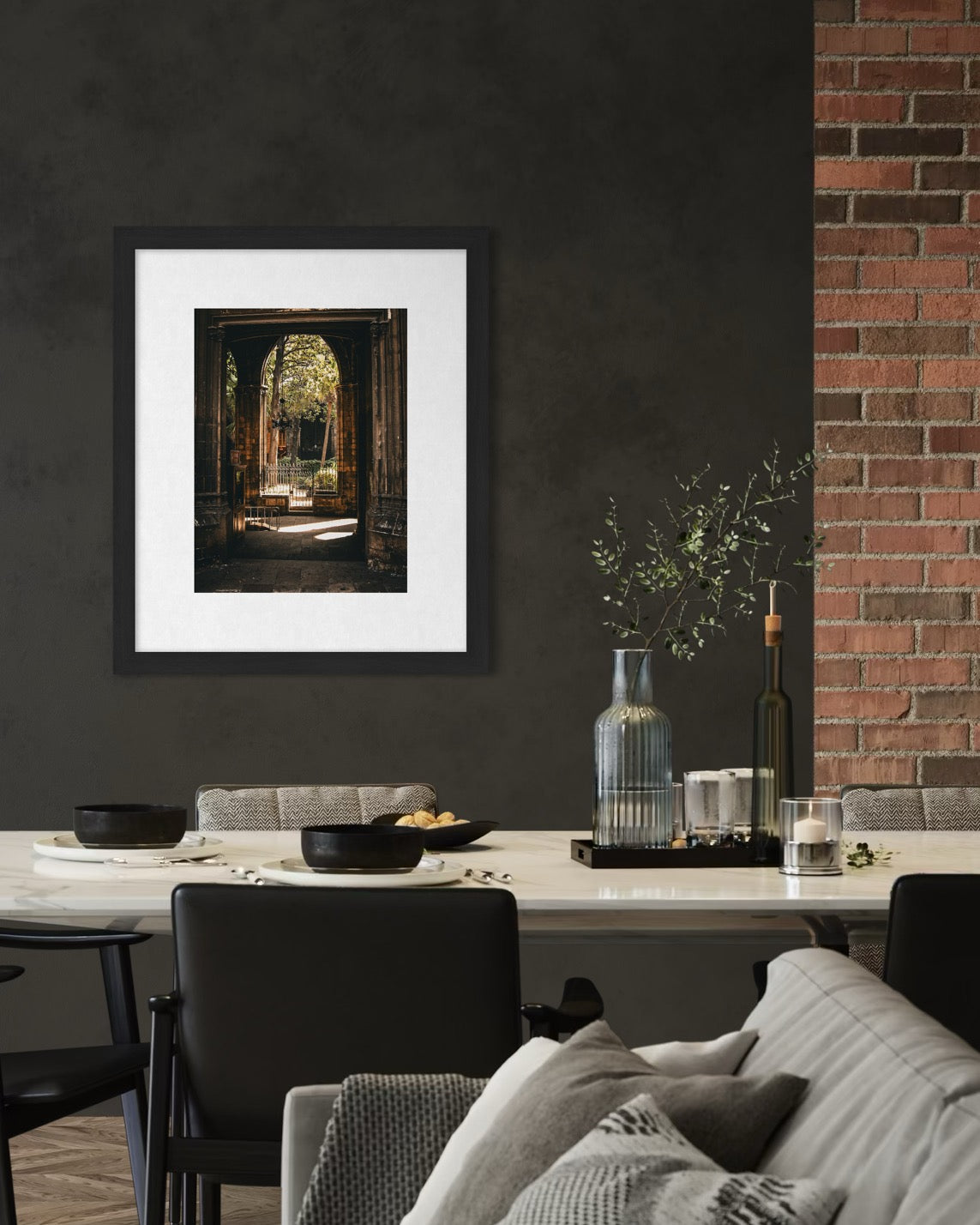 A modern dining area features a white table, black chairs, decorative vases, and "Barcelona Arch - Fine Art Print" by Leslie Brashear Photography. A section of exposed brick appears on the right.