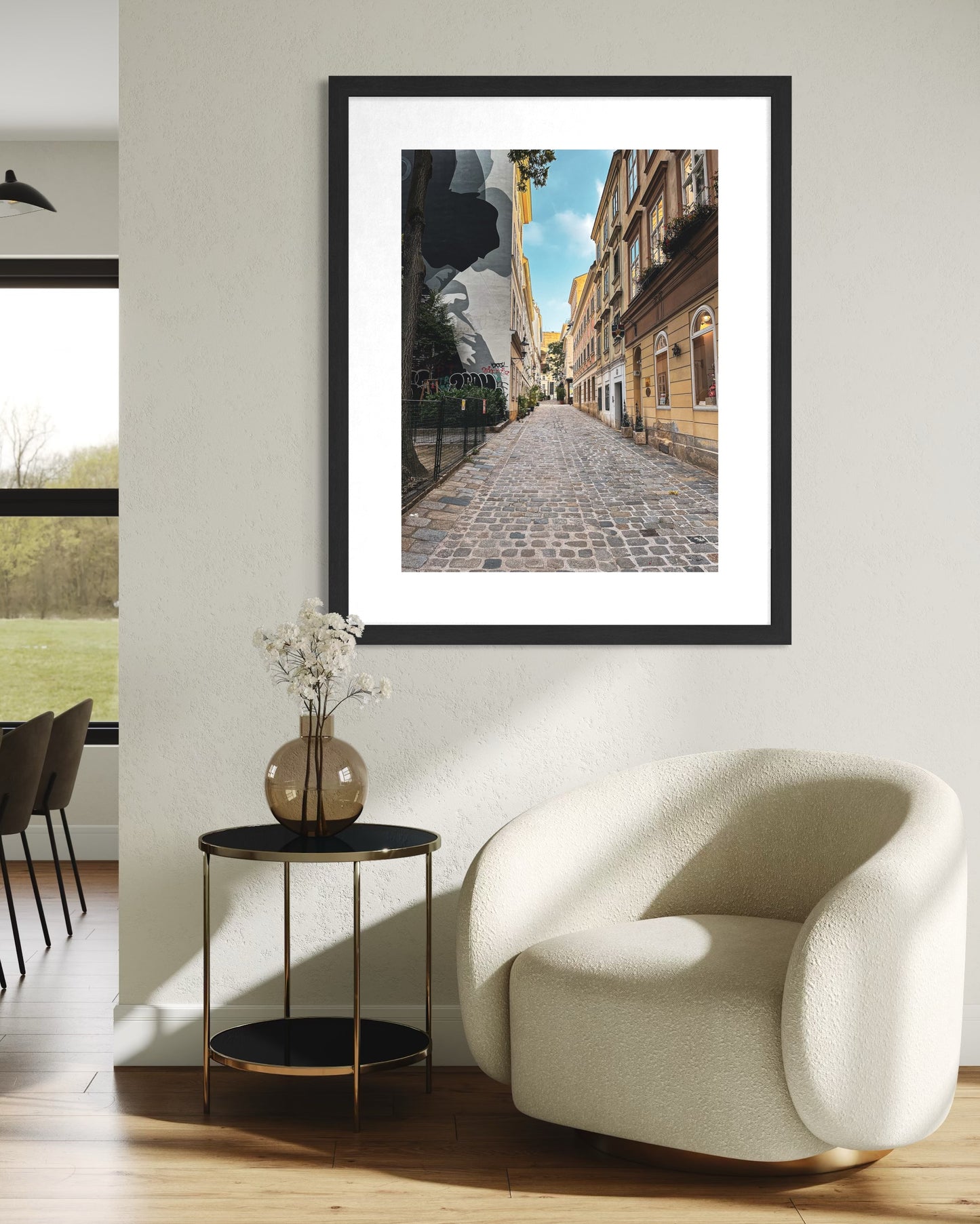 A modern living room features a white armchair, a round side table with a vase, and the “Quiet Alley in Vienna” fine art print by Leslie Brashear Photography displayed on the wall.