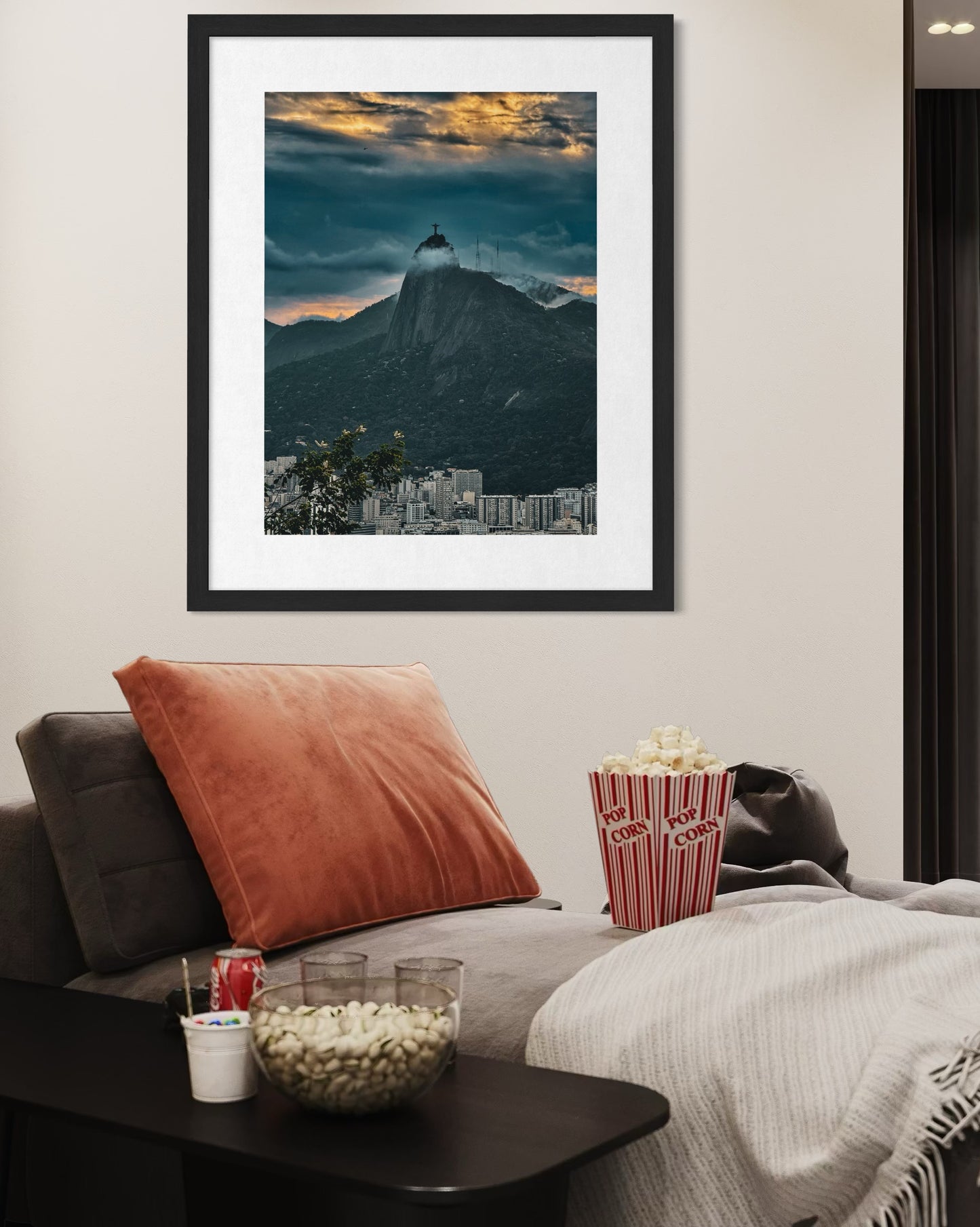 A Christ the Redeemer Amidst a Dramatic Sky - Fine Art Print by Leslie Brashear Photography hangs above a couch with popcorn, snacks, and a blanket in a stylish living room.