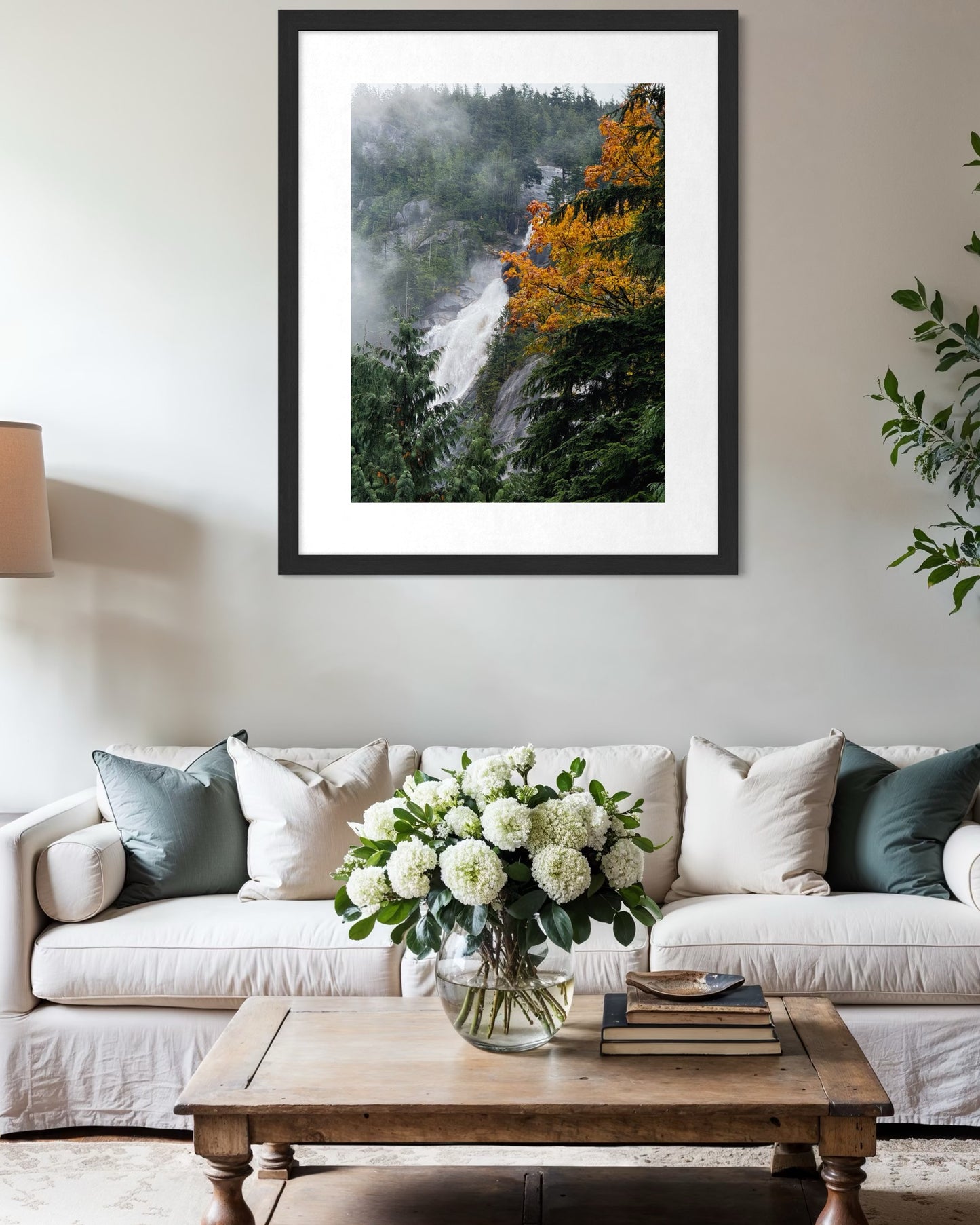 A living room with a white sofa, wooden coffee table, and glass vase of white flowers; above the sofa hangs "Autumn Cascade in the Mist" by Leslie Brashear Photography for a serene touch.