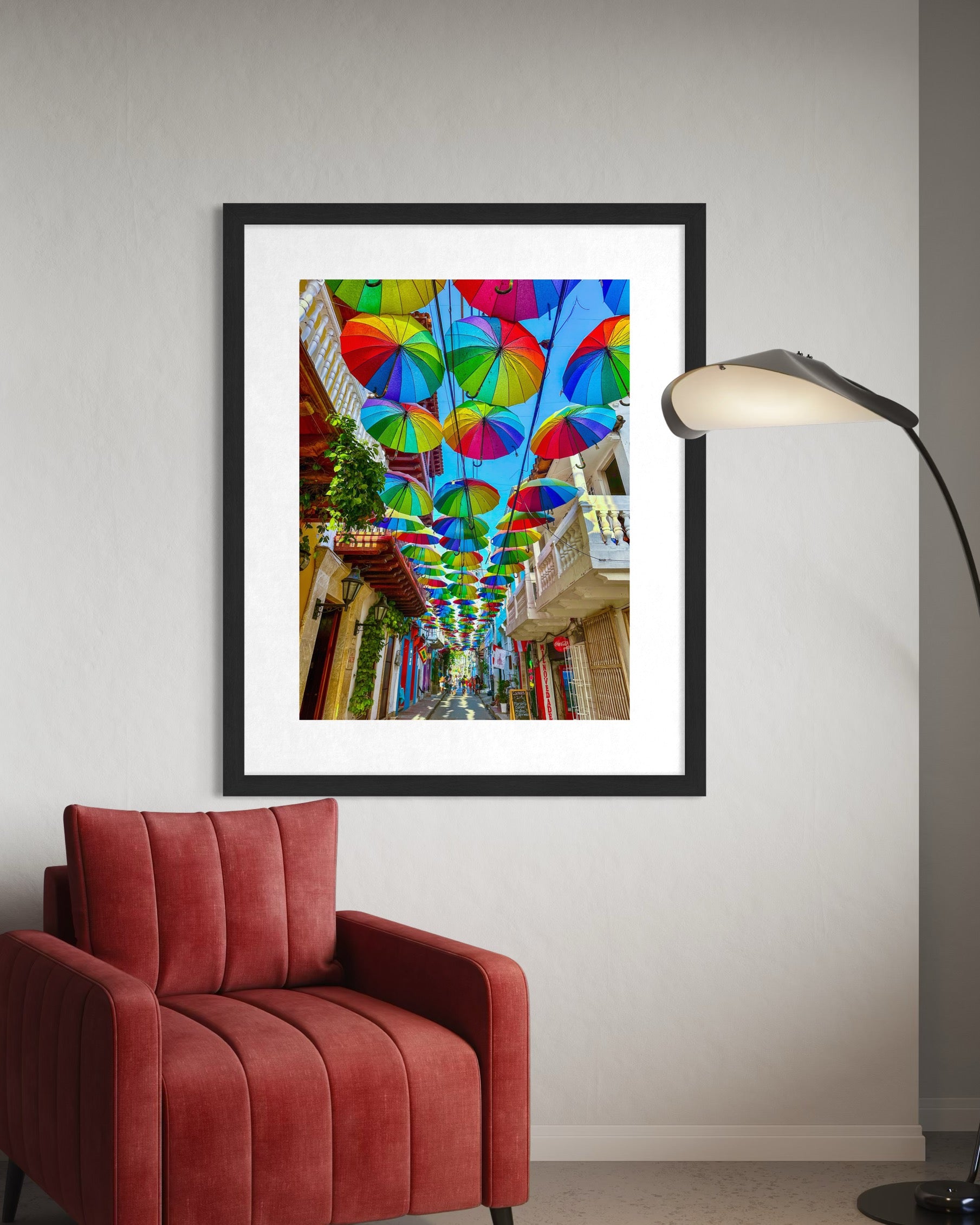 The "Umbrellas in Cartagena - Fine Art Print" by Leslie Brashear Photography features vibrant umbrellas above a street, shown framed on a white wall above a red chair and beside a modern floor lamp.