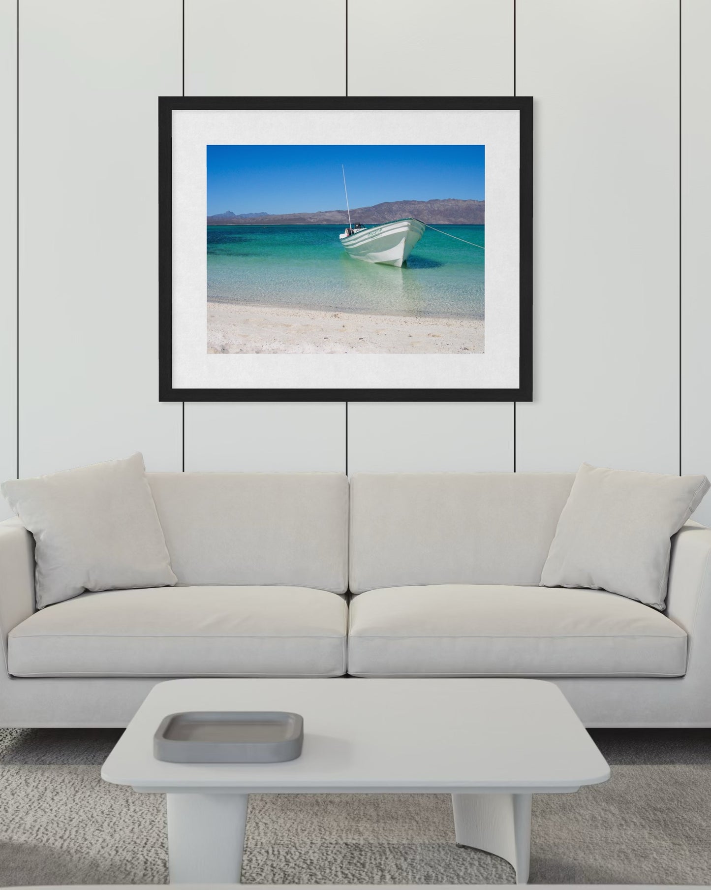 A white sofa and table in a modern living room, with "Peaceful Boat - Fine Art Print" by Leslie Brashear Photography—a beach and blue water scene from Loreto, Mexico—evoking the minimalist Sea of Cortez landscape, hanging above.