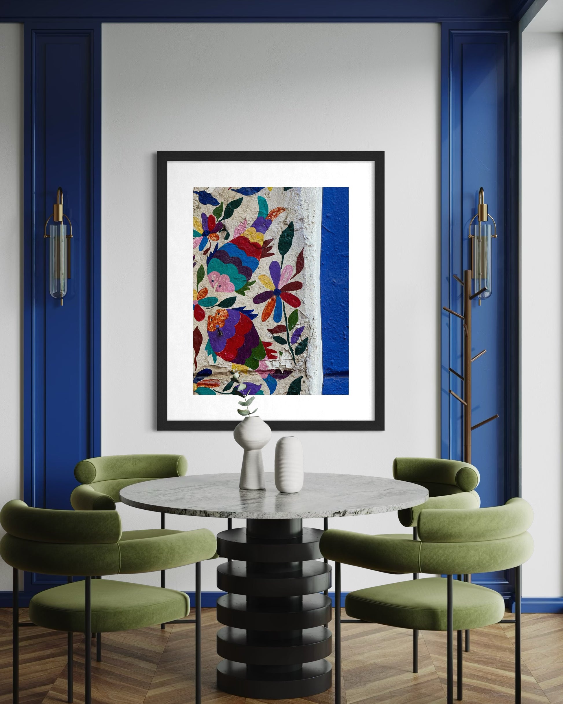 A modern dining area with green chairs and a round table, featuring the Vibrant Wall Art in Puebla fine art print by Leslie Brashear Photography, showcasing colorful Mexican floral embroidery on a white wall with blue trim.