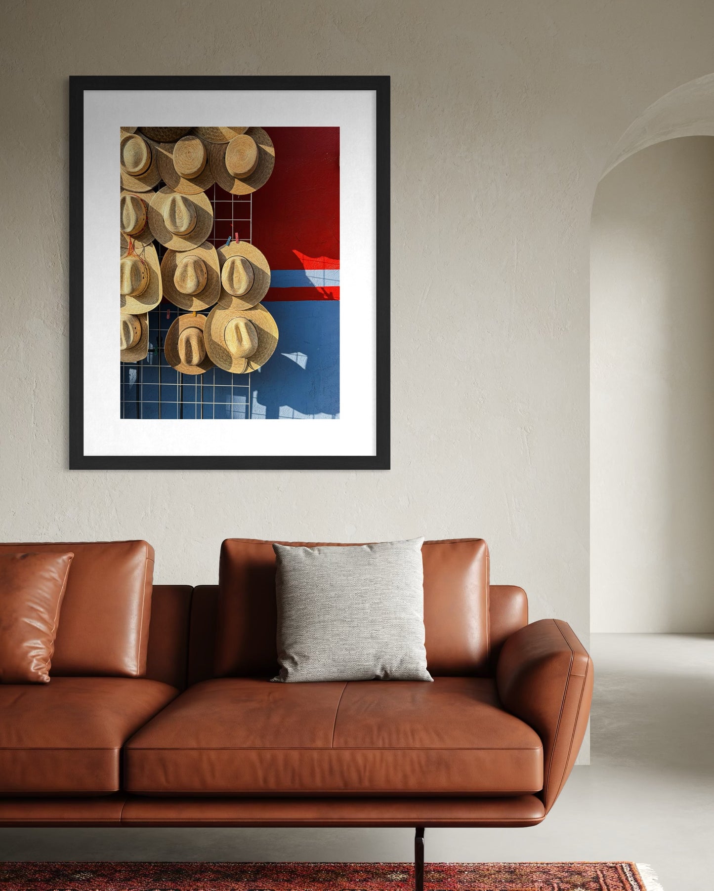 The "Straw Hats - Fine Art Print" by Leslie Brashear Photography features hats on a red and blue background, displayed above a brown leather sofa with a gray cushion—ideal Mexican marketplace wall art for your home.