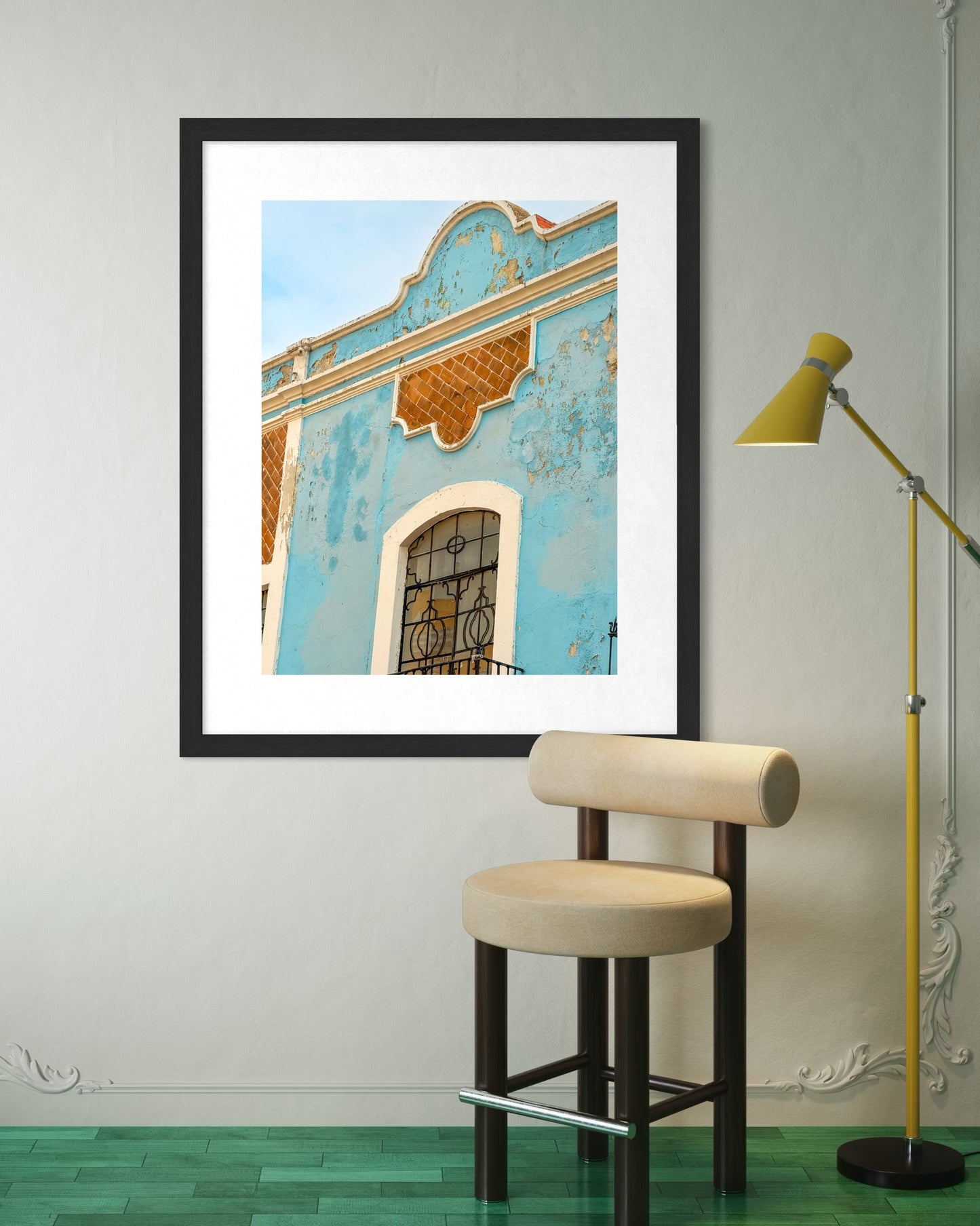 The "Blue Facade in Puebla - Fine Art Print" by Leslie Brashear Photography hangs above a chair and next to a yellow floor lamp on green flooring, featuring a weathered blue building facade with a decorative window.