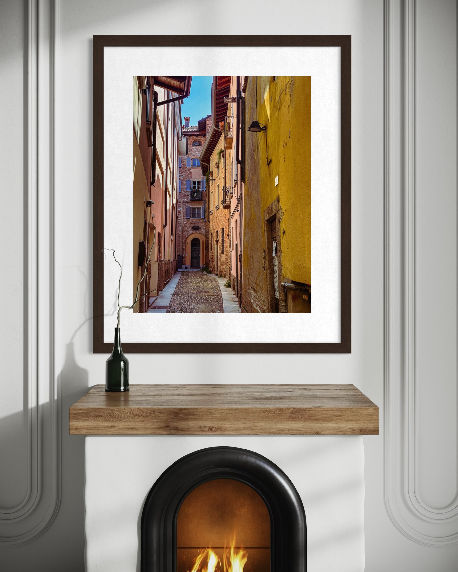Alleyway in Pavia - Fine Art Print by Leslie Brashear Photography captures a cobblestone street and rustic Italian architecture, beautifully displayed above a fireplace on a wooden mantel with a dark glass bottle.