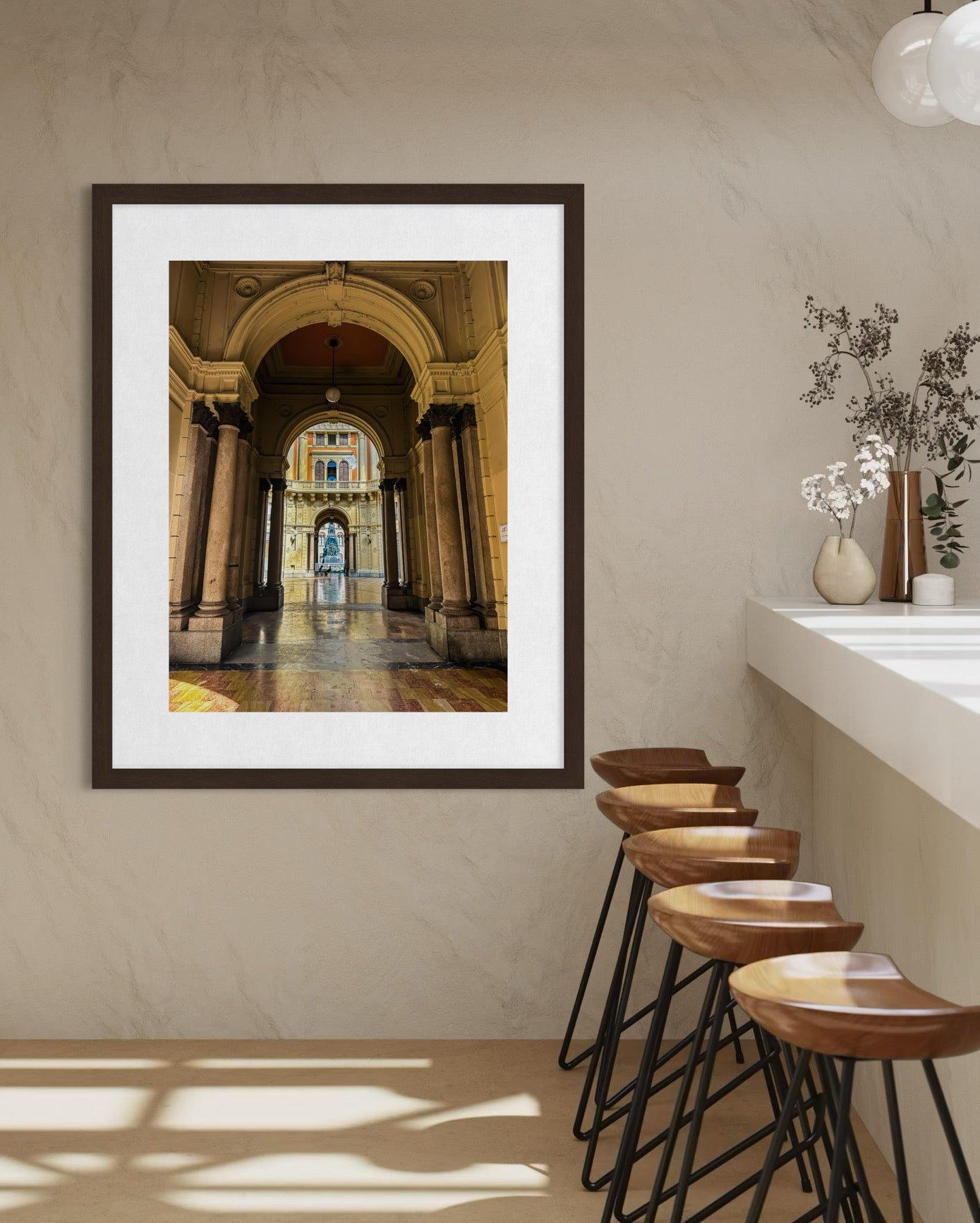 The "Timeless Arches of Pavia" fine art print by Leslie Brashear Photography hangs on a beige wall above wooden bar stools, next to a white counter topped with a vase of dried flowers.