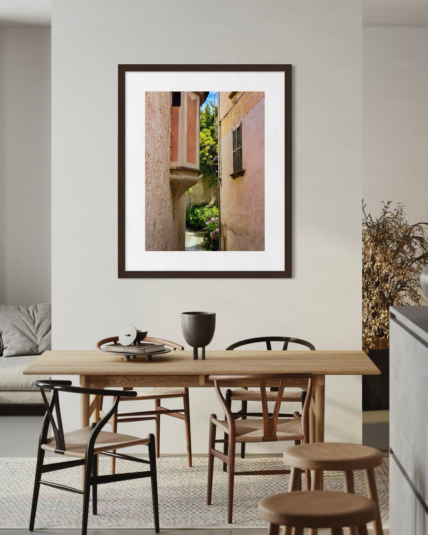 “Hidden Passage on Lago di Maggiore” fine art print by Leslie Brashear Photography, depicting a narrow alley with stucco buildings and greenery, hangs above a light wood dining table in a modern dining area.